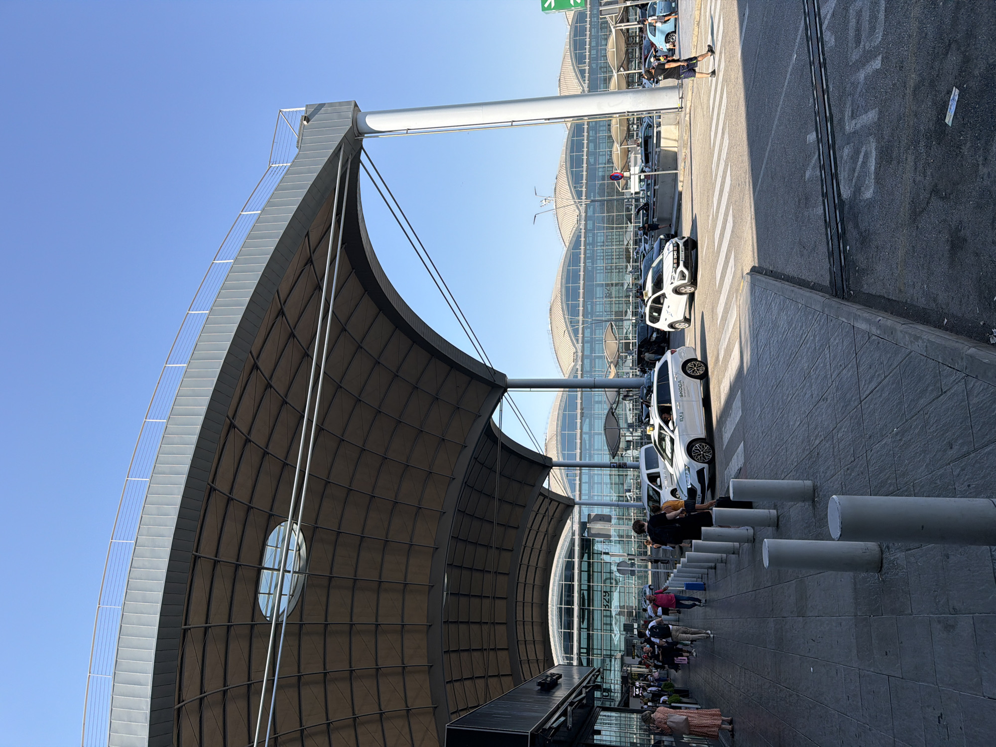 The image shows the exterior of Alicante-Elche Airport (ALC) in Elx / Elche, Spain. A large, modern terminal building with a distinctive curved roof structure dominates the scene. People are seen walking on the sidewalk outside the terminal, with several cars and taxis parked in front of the building. The sky is clear and blue. The architecture is characterized by its expansive glass facade and the prominent, wave-like canopy extending over the passenger pickup/drop-off area. The overall impression is one of a busy, contemporary transportation hub.