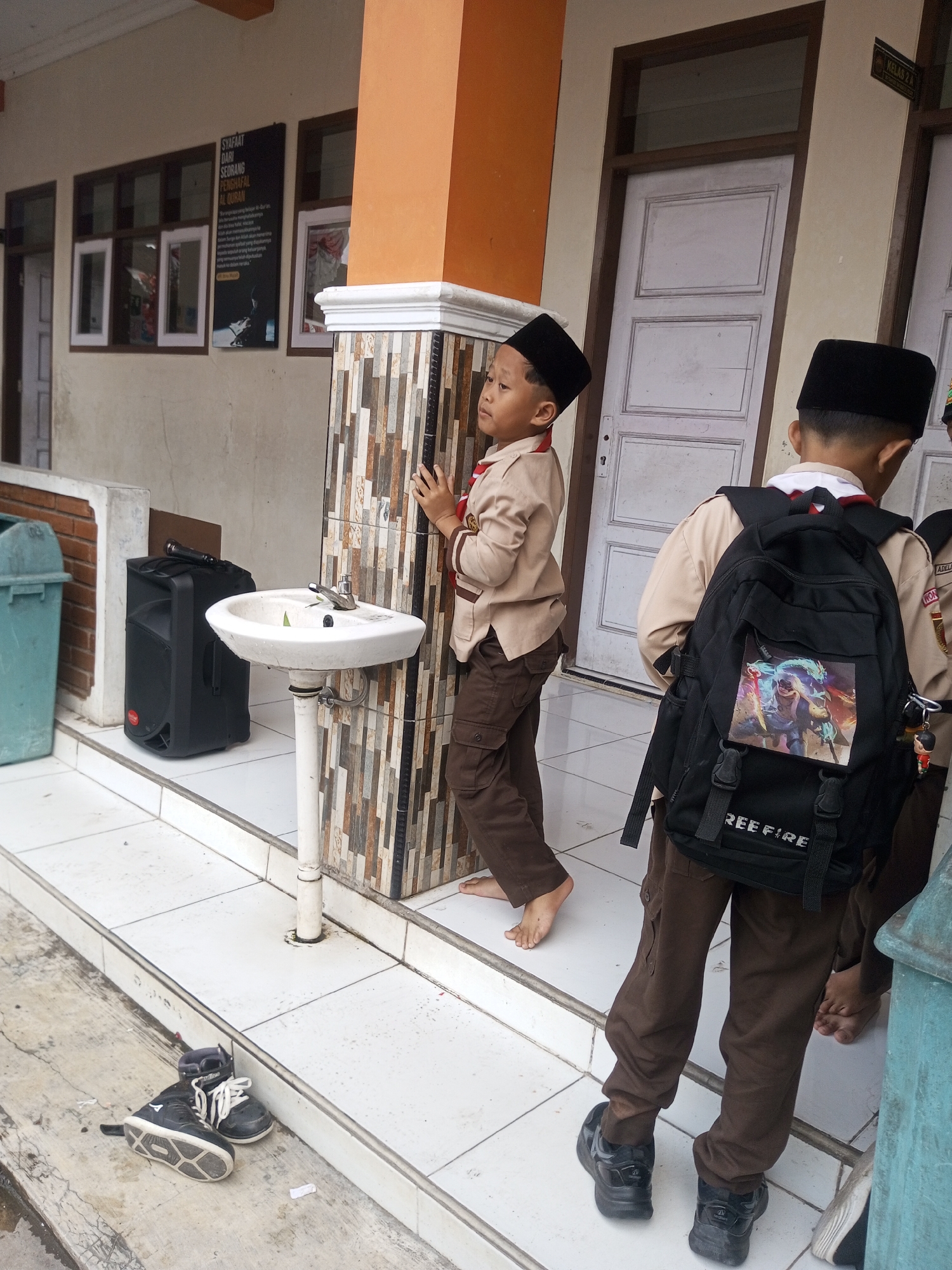 Two young boys, likely students, stand outside a building in Kalibeber, Indonesia. Both are wearing tan uniforms with brown pants and black peci hats. The boy on the left is facing left, looking away from the camera, with his right hand touching a tiled column. He is barefoot. The boy on the right has a black backpack with a "FREE FIRE" logo and a picture of a video game character on it. He is facing away from the camera, and his left foot is visible. Nearby, there is a white sink with a faucet, a black speaker with a microphone on top, and a light blue trash can. A pair of black and white sneakers are placed on the ground in the foreground. The building has white doors with dark frames and windows, and an orange-colored section above. The ground appears to be tiled and concrete.