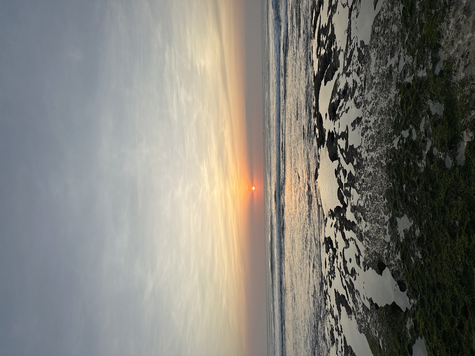 This image depicts a sunset over the ocean in Kuta Selatan, Indonesia. The sun, a vibrant orange orb, is setting just above the horizon, casting a warm glow across the sky. The sky is a gradient of soft oranges, pinks, and pale blues, with wispy clouds scattered throughout. Gentle waves roll towards the shore, creating a serene atmosphere. In the foreground, the rocky coastline is exposed, with tide pools reflecting the colorful sky. Patches of green algae cover some of the rocks, adding a touch of natural texture. The overall scene is peaceful and picturesque, capturing the beauty of a tropical sunset.