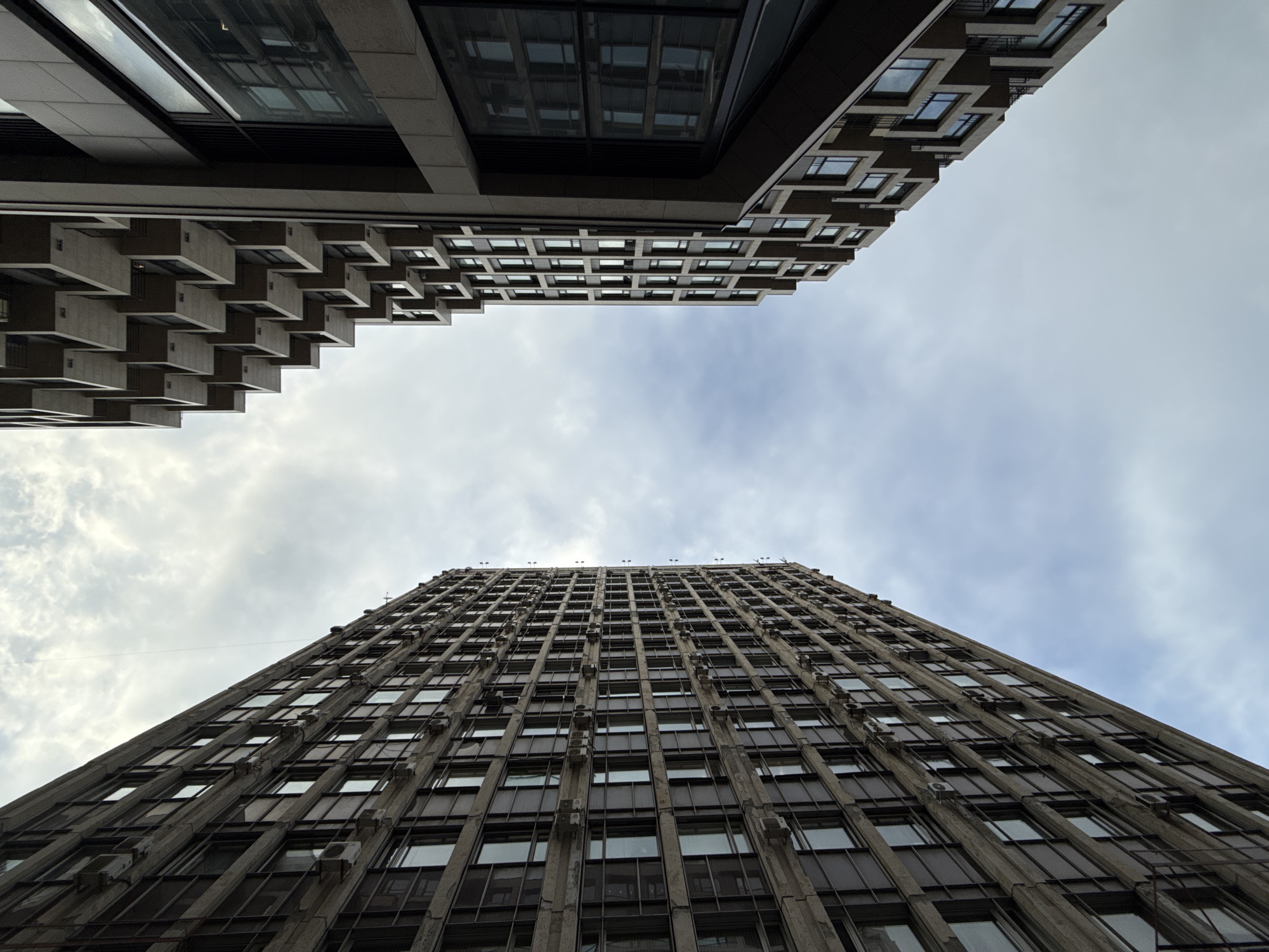 The image shows a worm's eye view of two buildings against a cloudy sky. The building at the bottom is tall and slender, with a repeating pattern of windows and what appears to be air conditioning units. The upper building has a more modern design with cubic protrusions. The photograph was taken in Moscow, Russia.