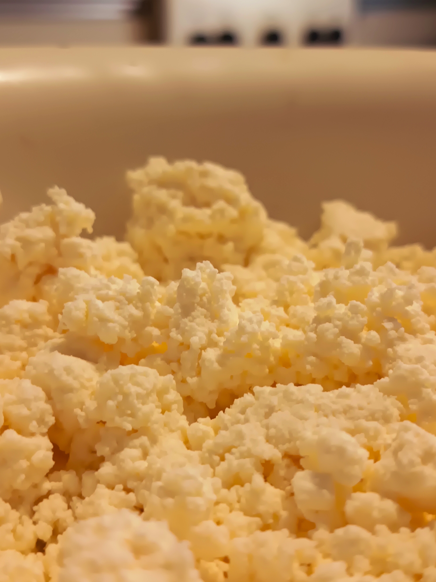 This is a close-up, eye-level, indoor photograph of a white, lumpy substance that appears to be cottage cheese or a similar dairy product, filling the lower half of the frame. The substance is a pale yellow and white color with a textured, granular appearance. In the background, a blurred, out-of-focus beige surface is visible, with darker shapes that could be indistinct objects or shadows. The lighting is warm and appears to be artificial, casting soft shadows. There are no people, animals, or clear indications of a specific location within the city of Izmail, Ukraine. The image focuses solely on the food item and its immediate, indistinct surroundings.
