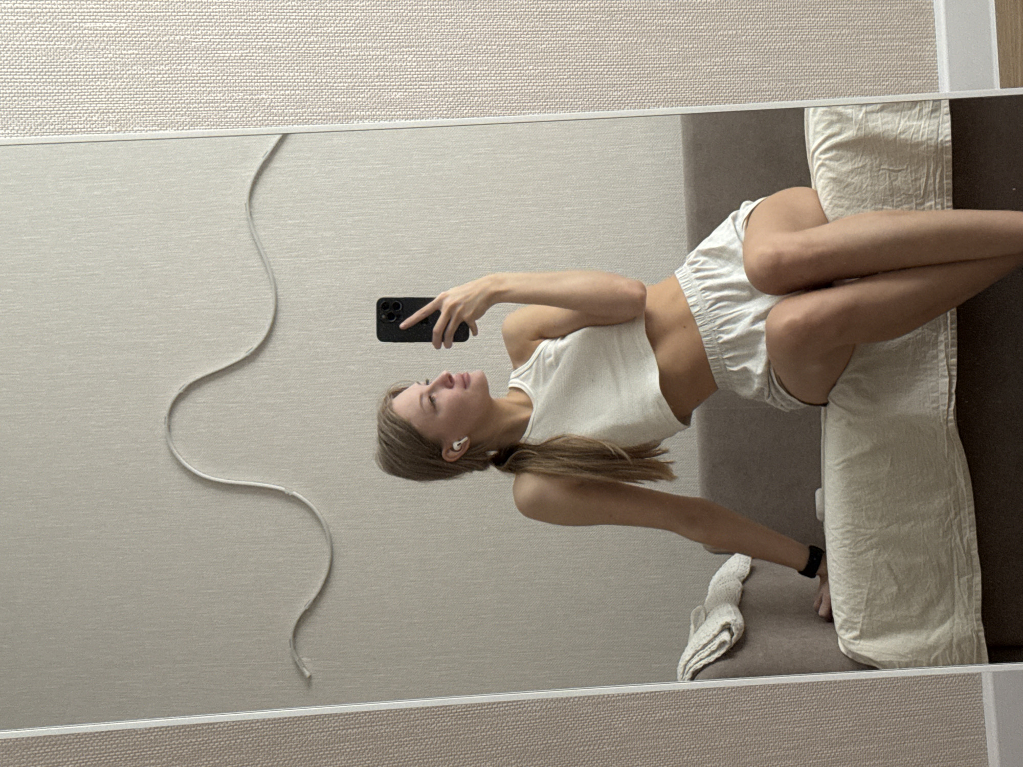 A young woman with blonde hair pulled into a ponytail takes a mirror selfie. She is wearing a white crop top and white shorts, and has white earbuds in her ears. She is sitting on a sofa with a beige textured wall behind her. There is a decorative wavy line on the wall. The woman is holding a black smartphone in her right hand to take the picture. A black fitness tracker is visible on her left wrist. A light-colored throw is draped over the arm of the sofa. The scene appears to be indoors, likely in a residential setting. The location is identified as the city of Perm, Russia.