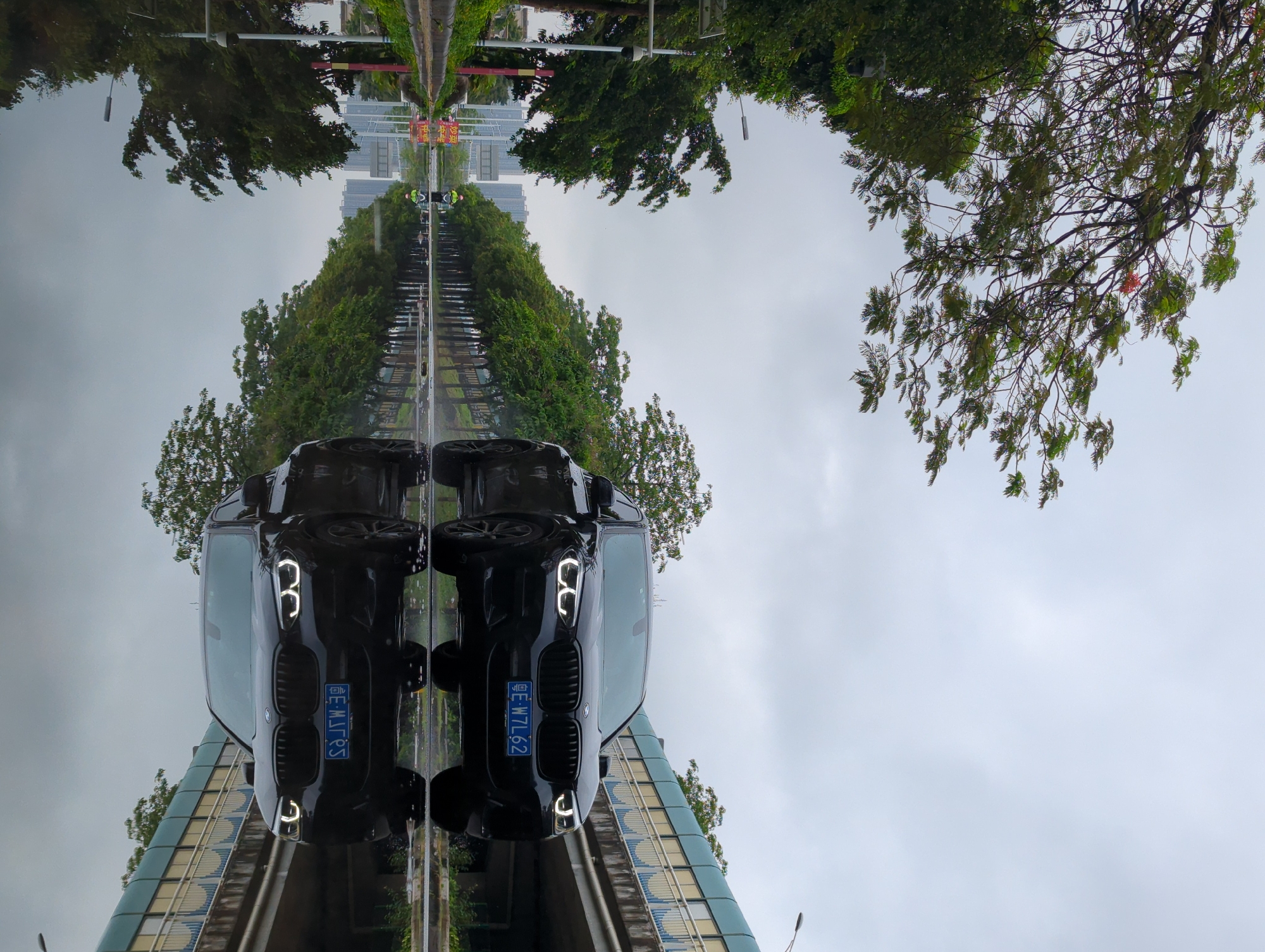 The image shows a reflection of a street scene in Shunde District, China, featuring two black BMW X vehicles parked side-by-side on a wet surface. The reflections include surrounding trees, buildings, and street signs. The license plate on the left car reads "粤E MJ7R5" and the license plate on the right car reads "粤E W7L62". The sky is overcast.