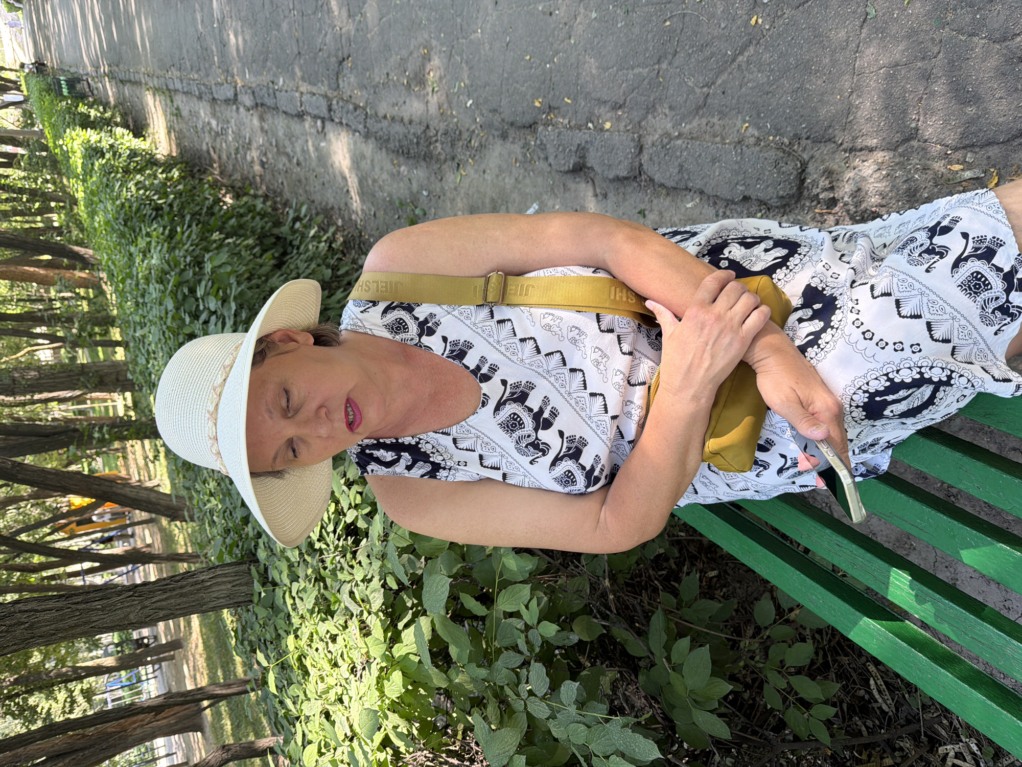 A white woman wearing a wide-brimmed hat and a white and black elephant-patterned dress sits on a green park bench. She is holding a smartphone in her hands and looking down at it. A yellow crossbody bag is slung over her right shoulder. The background shows a park with trees and a paved pathway.