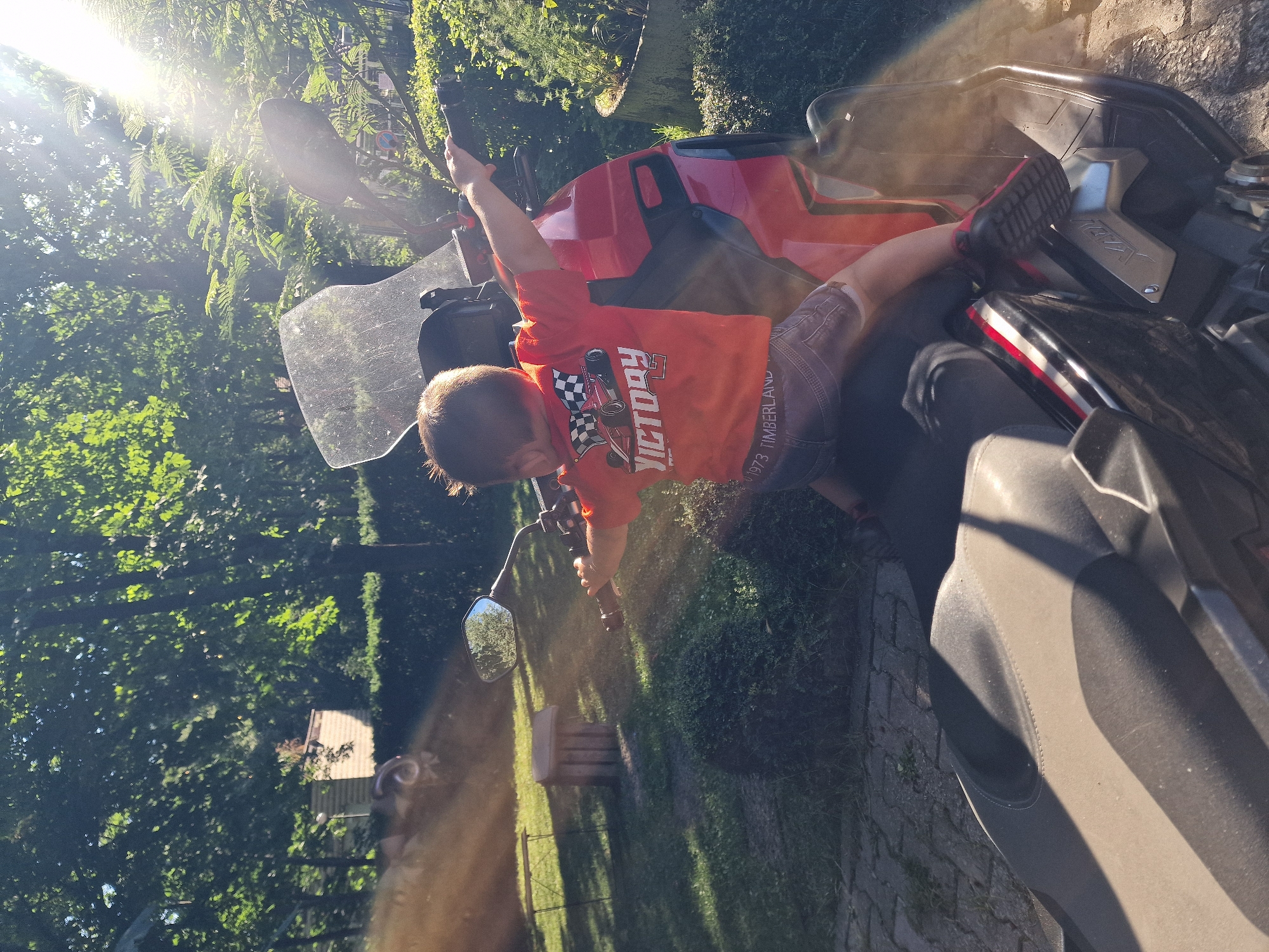 In an outdoor, sunny environment, a young child is seated on a red and black motorcycle. The child, viewed from behind, is wearing an orange t-shirt with a checkered flag and "VICTORY" graphic on the back, and denim shorts. Their arms are outstretched as if riding, and they are holding onto the handlebars. The motorcycle has a clear windshield. The background consists of lush green trees and foliage, with sunlight filtering through the leaves, creating lens flare effects in the image. A portion of a seat is visible in the foreground.