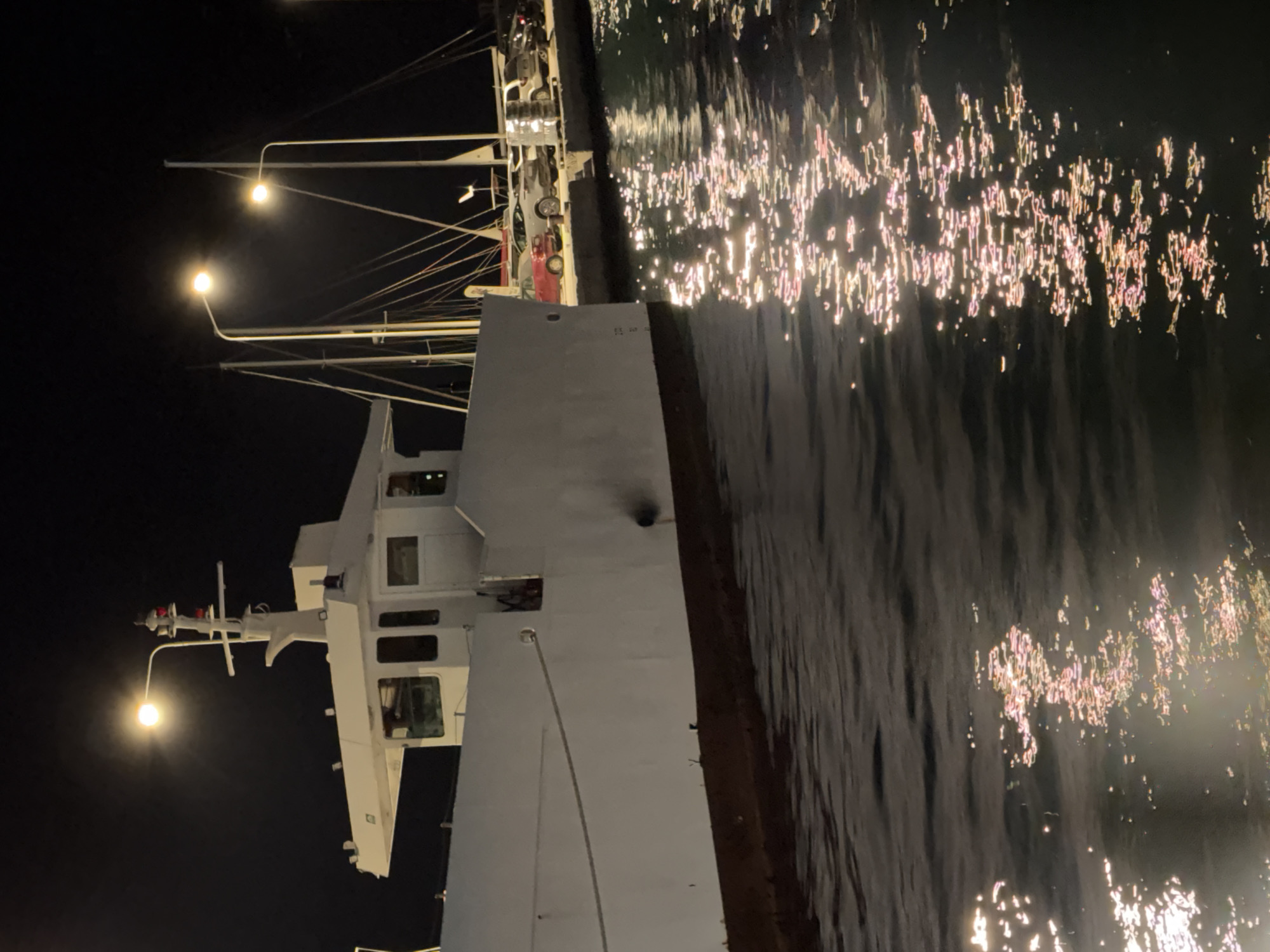 The image depicts a night scene at a harbor in the Municipal Unit of Alyzia, Greece. A white ferryboat is docked, its hull partially submerged in dark, rippling water. The water reflects the lights from the shore, creating a shimmering, pinkish-gold pattern.
On the shore, several streetlights illuminate the area. Behind the ferryboat, a row of parked cars is visible, including a red sedan and a silver SUV. The background shows the masts of sailboats and a dark, starless sky. The overall impression is one of a quiet, dimly lit port at night.