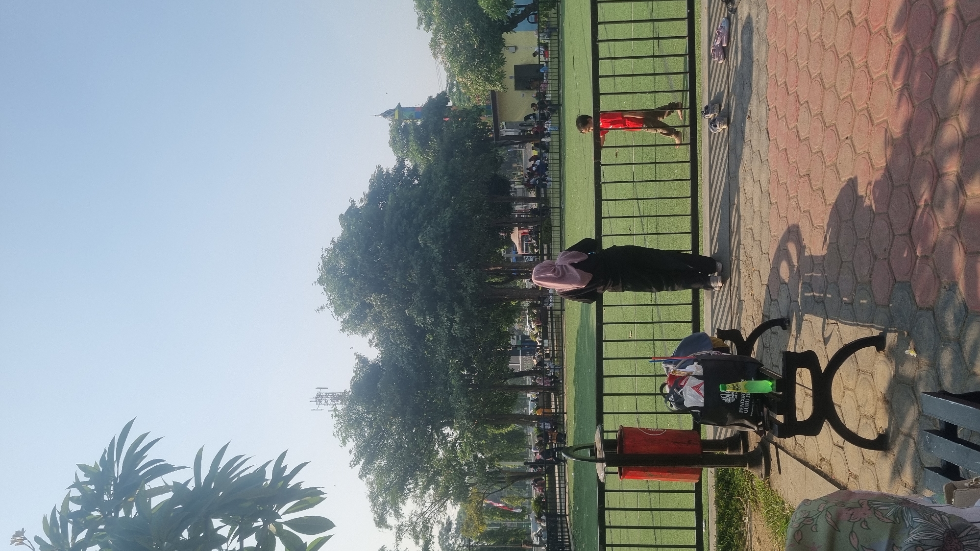 In Kaliwungu, Indonesia, an outdoor scene captures a park area with a focus on a green field enclosed by a black metal fence. A woman wearing a pink hijab and black clothing stands facing away from the camera, looking towards the field. A young boy in a red shirt and brown pants is also on the field, walking away from the woman.
In the foreground, a black bench holds a collection of items including a bag with a bottle of green soda on top. To the left of the bench is a red trash can. The ground is paved with hexagonal tiles in shades of red, grey, and tan, with shadows cast across it.
In the background, numerous trees provide shade, and further beyond, a distant tower and a colorful building with a spire are visible. Other people are scattered in the background, seated and standing, suggesting a public gathering space. The sky is clear and blue.