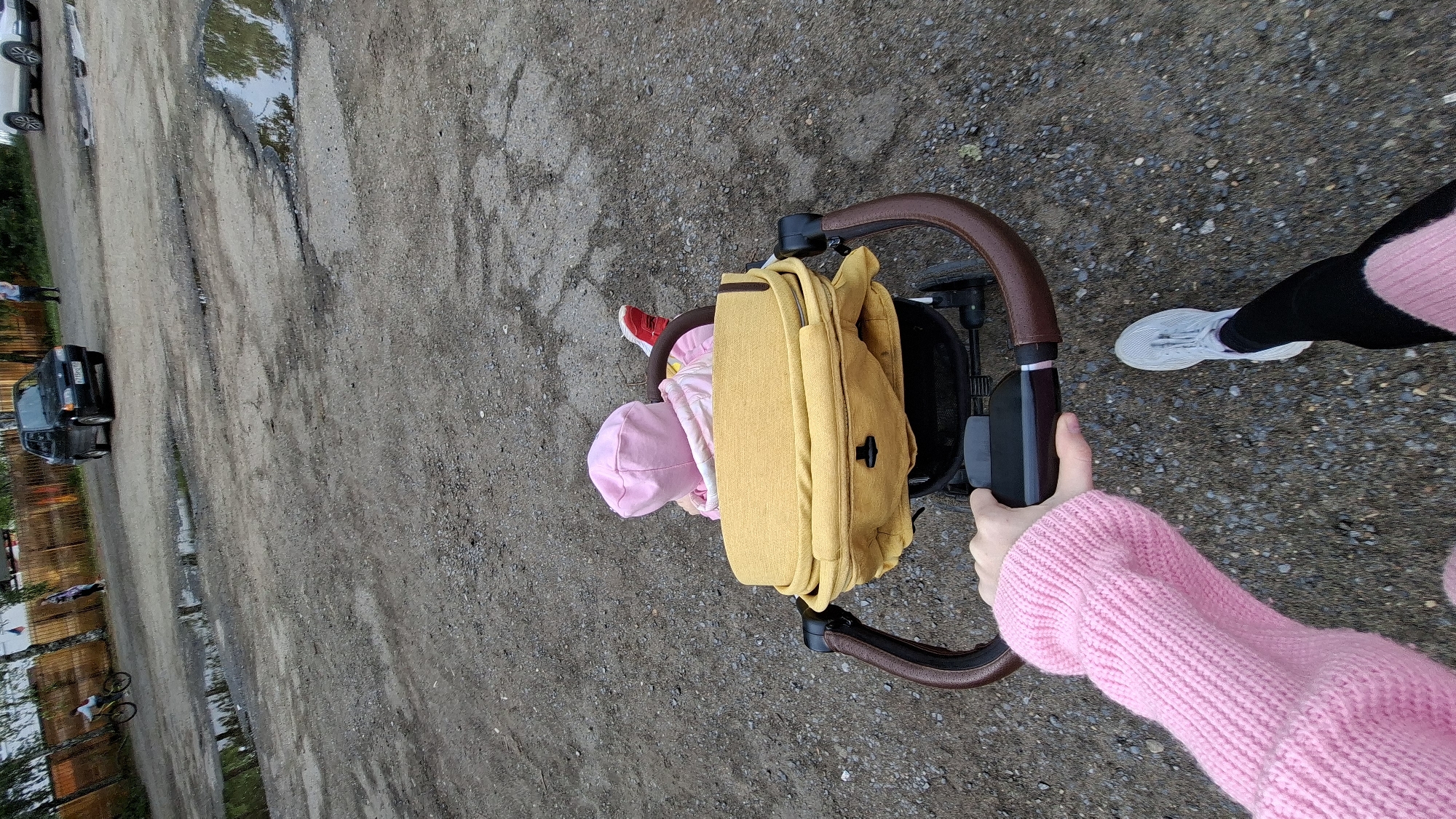 An overhead shot captures a person pushing a stroller along an unpaved, gravelly road in Chaikovsky, Russia. The person pushing the stroller is partially visible on the right, wearing a textured pink knitted sweater and black leggings with white/light grey sneakers. Their hand grips the brown-handled stroller.
Inside the stroller, a small child is visible from the back, wearing a pink hooded top or hat, with a flash of a red shoe. A mustard yellow bag or backpack is attached to the back of the stroller, partially obscuring the child. The stroller features a black frame and a brown, curved handle.
The ground is a mix of gravel and dirt, showing signs of wear with several potholes and puddles reflecting the sky and some distant trees. In the background on the left, several vehicles are parked or driving. A dark-colored car, possibly an older model like a Lada, is visible further down the road, and a white car is partially seen behind it. Further left, a wooden fence lines what appears to be a property, and a bicycle is parked near it. The overall impression is of a casual walk on a somewhat rustic road, possibly after rain given the puddles. No legible text is visible in the image.