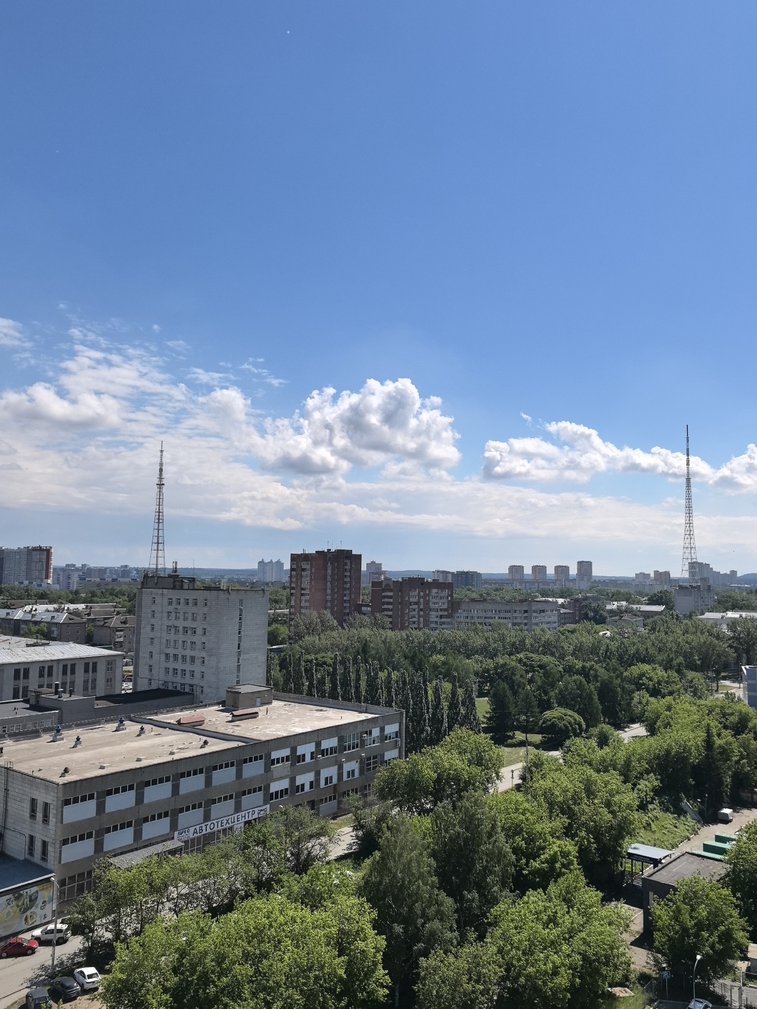 The image shows a daytime aerial view of a cityscape in Perm, Russia. The sky is predominantly blue with scattered white clouds. In the foreground, there are several concrete buildings, the largest of which has the Russian word "АВТОТЕХЦЕНТР" visible on its facade. Numerous trees cover a significant portion of the middle ground, interspersed with roads and smaller buildings. In the background, there are apartment buildings and other urban structures, with two prominent radio towers. The overall scene suggests a typical urban landscape in Perm, Russia.