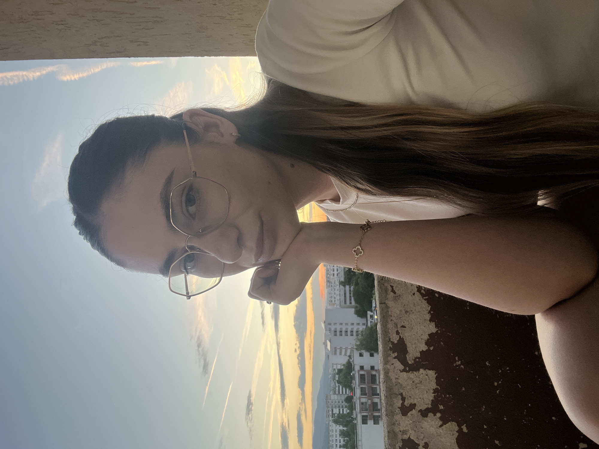 A young woman with long brown hair and glasses is posing for a selfie on a balcony with a city skyline in the background during sunset. The woman is wearing a white t-shirt and a bracelet with a four-leaf clover charm. The city appears to be dense with apartment buildings. The sky is a mix of blue and orange, with some clouds.