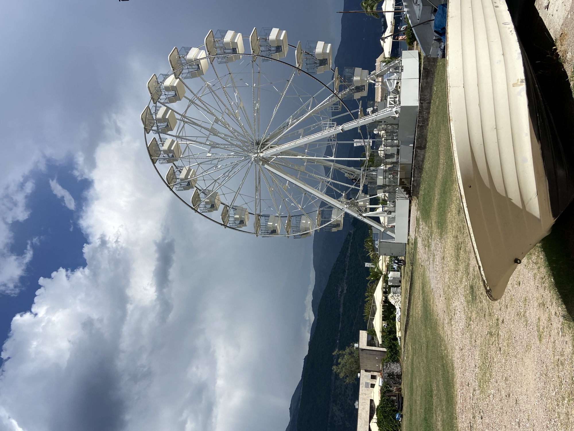 The image displays a Ferris wheel set against a backdrop of mountains and a cloudy sky. The Ferris wheel is white and appears to be a tourist attraction. In the foreground, a light-colored boat is positioned on the right side of the frame, taking up a significant portion of the image. A grassy area with some gravel is visible in front of the boat, and further beyond that, there are buildings and palm trees, suggesting a resort or coastal town setting. The sky is partly cloudy, with some areas of dark clouds, indicating potentially changing weather. The mountains in the background are covered in dense vegetation and appear to be quite large. There is no visible text in the image. The scene seems to be captured during the daytime, with the lighting suggesting it could be mid-morning or afternoon. The overall atmosphere appears calm and scenic, with the focus on the juxtaposition of the amusement park ride with the natural landscape and the man-made boat.