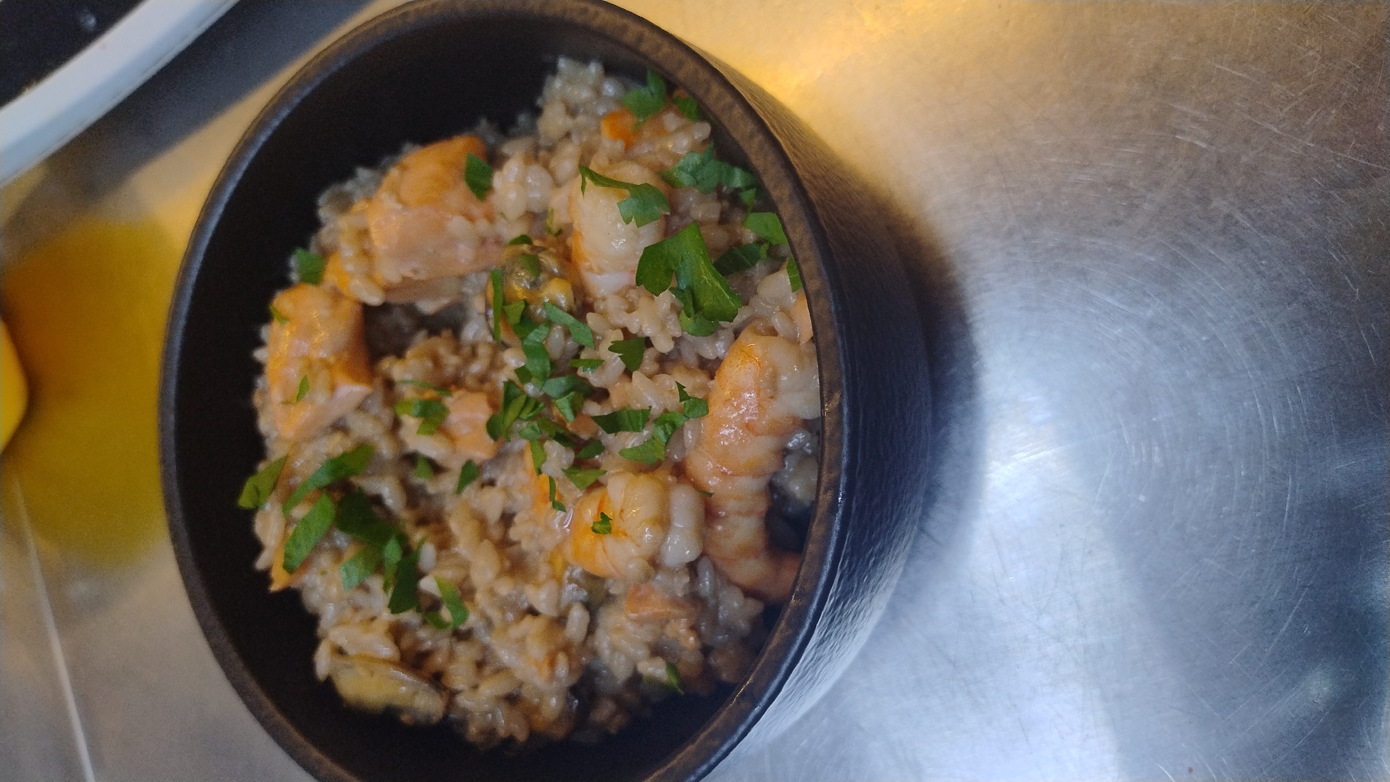 A close-up, overhead shot shows a dark-rimmed bowl filled with seafood risotto. The risotto contains rice, shrimp, and pieces of salmon, all garnished with chopped parsley. The bowl sits on a brushed metal surface. The background is blurred, with hints of yellow and white. The image was taken in the city of Mykolaiv, Ukraine.