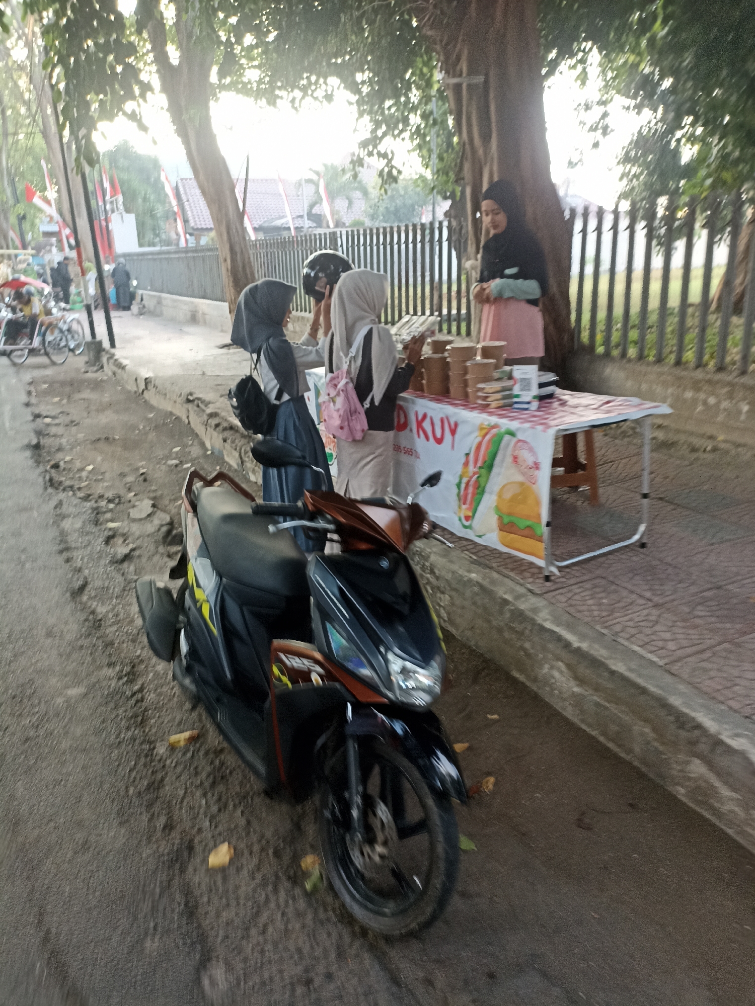 In Mojopanggung, Indonesia, a scene unfolds beside a tree-lined street, where a motorcycle is parked on the roadside. To the right, a white table is set up, displaying several food containers and a QR code. A woman in a black hijab and a pink dress stands behind the table, seemingly attending to the stall. Two other individuals, also in hijabs, are visible near the table. In the background, a fence lines the street, and red and white flags are displayed on poles. Further back, several bicycles are parked. The lighting suggests it is either early morning or late afternoon, casting a warm glow on the scene.