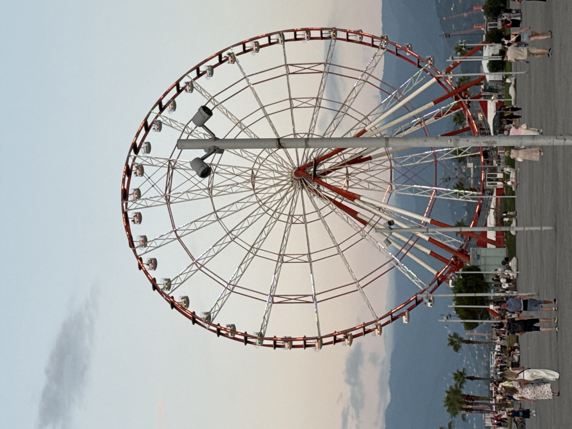 The image shows a large Ferris wheel, a popular amusement park ride, with a red and white structure. It is situated in an open area, possibly a promenade or park, with a crowd of people visible in the background. Palm trees and distant mountains are also part of the scenery, suggesting an outdoor recreational location. The sky is a soft, muted color, indicating it might be evening or dusk. The Ferris wheel appears to be illuminated with decorative lights, some of which are visible on its rim. In the foreground, a gray pole with two lights is prominently positioned. People are seen walking and standing, some observing the Ferris wheel or engaging in activities typical of a public gathering space.