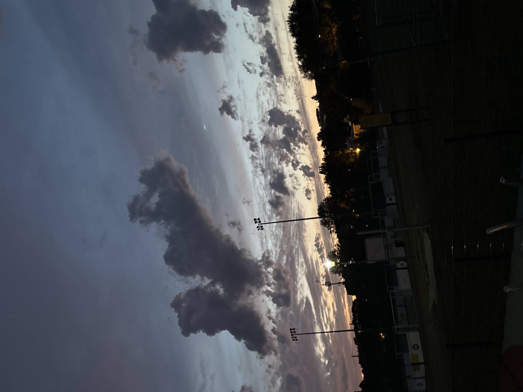 The image shows a twilight scene over a sports field, likely a futsal or small soccer pitch, in Lublin, Poland. The sky is filled with dramatic, dark clouds illuminated by the setting sun, casting hues of purple, orange, and blue. Two tall light poles with multiple lamps are visible on the field, suggesting it is used for evening activities. The field itself is dark, with outlines of the goals and surrounding fences visible. Trees line the background, and some buildings are faintly illuminated in the distance. The overall impression is one of a quiet, atmospheric evening in an urban park or recreational area.