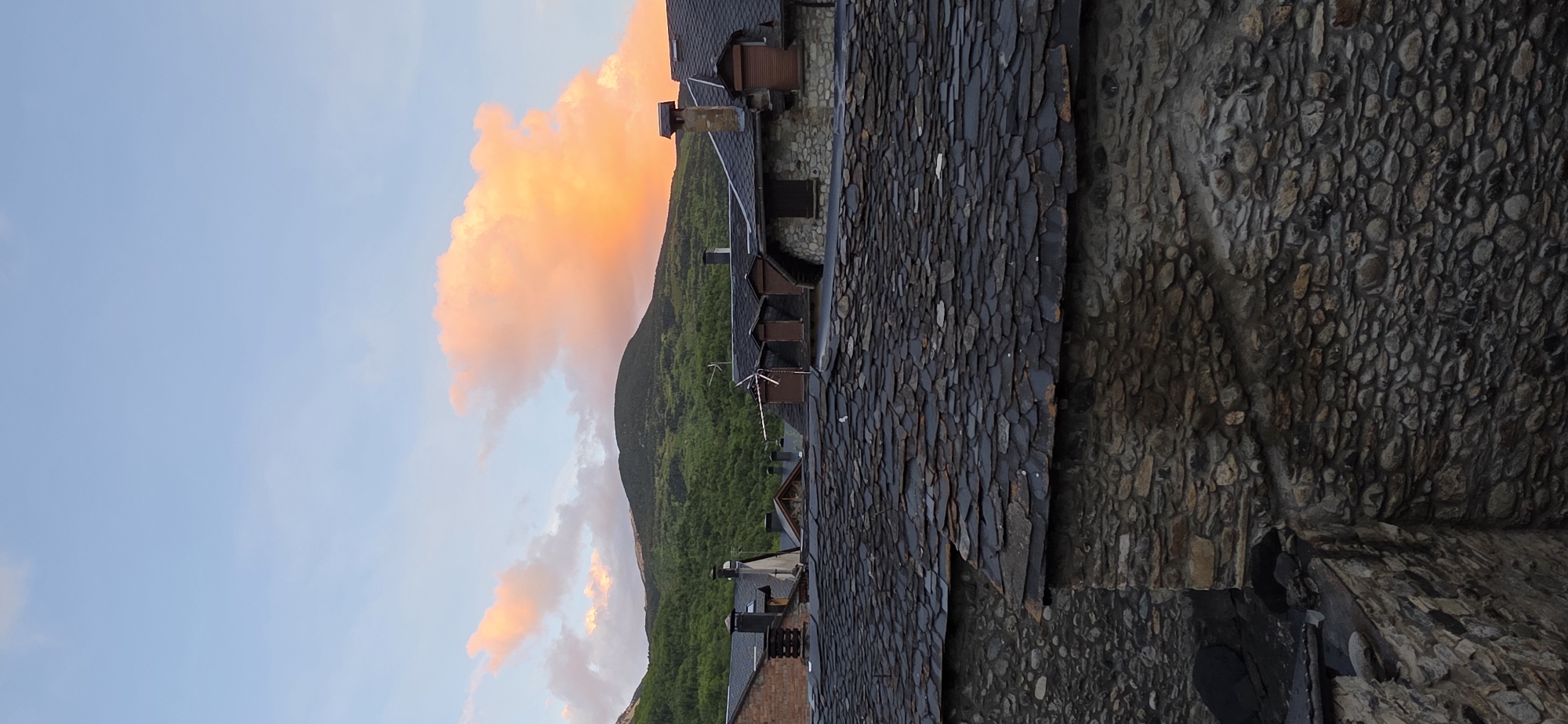 The image shows a view in the town of Benasque, Spain. In the background, a mountain covered in green vegetation is seen. A cloud tinged with orange and pink due to the setting sun is present in the sky. In the foreground, the gray-tiled roof of a building is prominently displayed. A section of a stone wall is also visible, constructed with rounded stones. The perspective suggests the photo was taken from a higher vantage point, possibly another building.