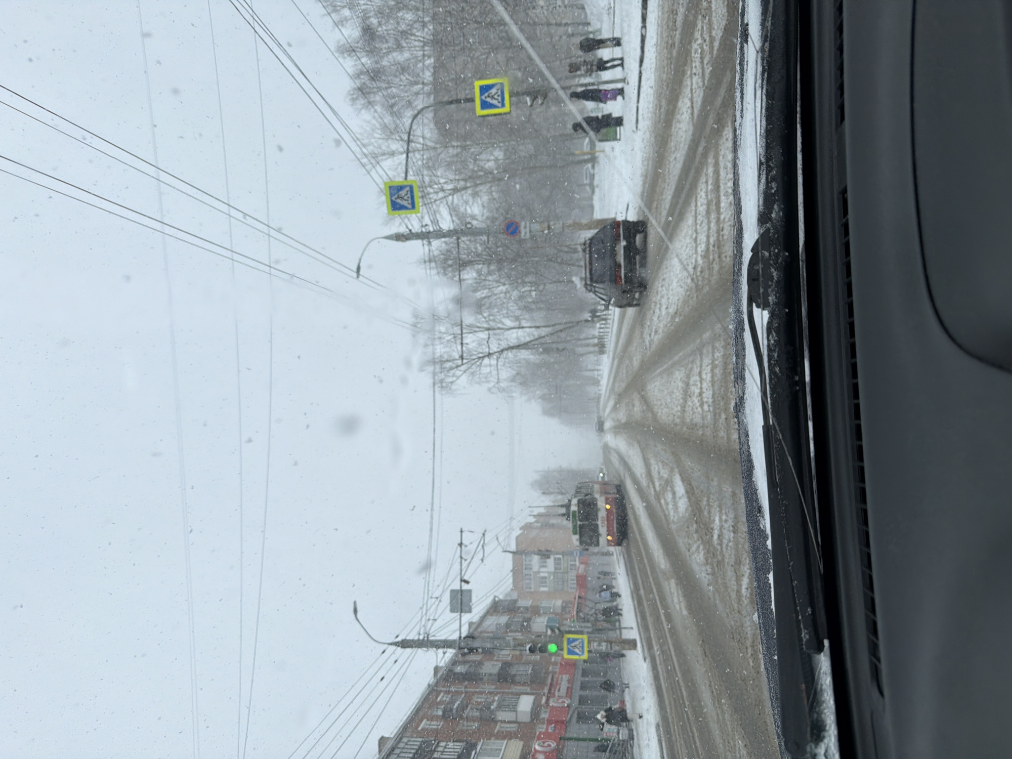 The image depicts a snow-covered street scene in Izhevsk, Russia, viewed from inside a vehicle. The road is covered in slush and tire tracks. Several vehicles are visible, including cars and what appears to be a trolleybus. Buildings line the street, and overhead electrical wires are present. Pedestrian crossing signs are visible, and a few people can be seen walking on the sidewalk. The scene is overcast with falling snow.