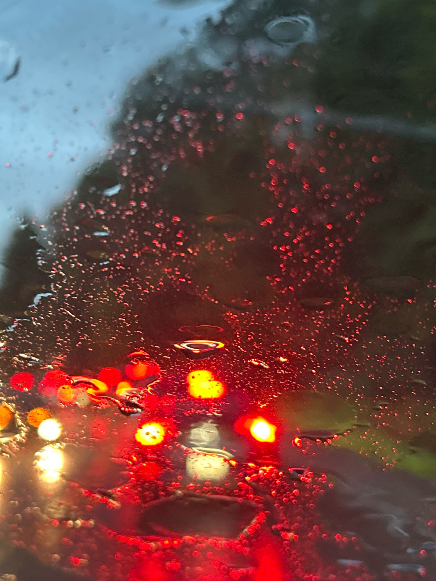 A close-up, low-angle shot captures a blurred view through a rain-streaked windshield, depicting a traffic scene at dusk. Numerous red and yellow lights, indicative of taillights and traffic signals, are scattered across the frame, softened by the water droplets on the glass. The background is a dark, indistinct blur of trees and sky. The image was taken in Upper St. Clair, United States of America.
