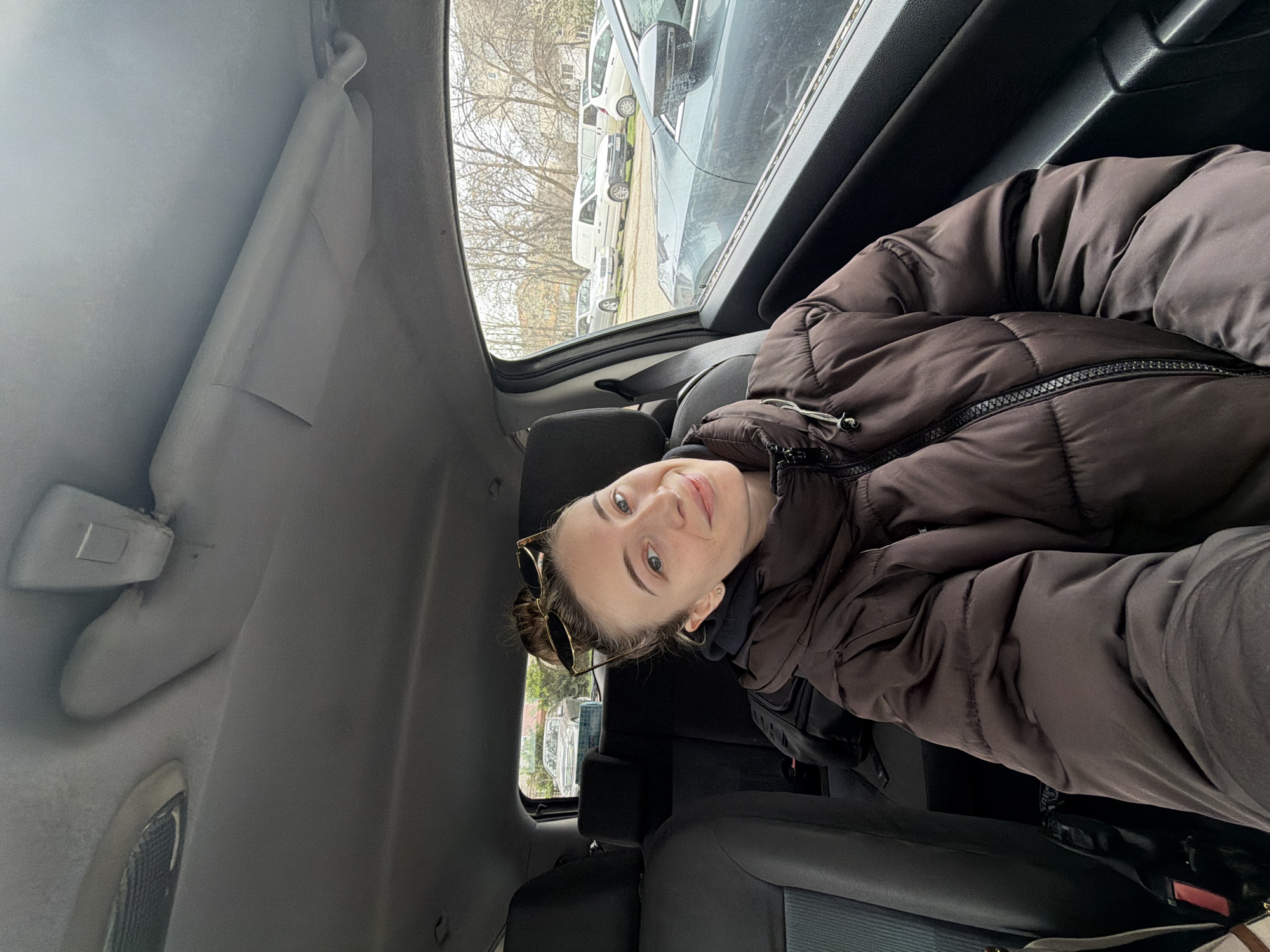 A woman is sitting in the back seat of a car. She is wearing a brown puffer jacket. Sunglasses are perched on top of her head, which has a bun hairstyle. The car appears to be parked on a street; several other cars are visible through the window along with leafless trees and a building. The location appears to be in Plovdiv, Bulgaria. The interior of the car is grey. The woman is looking at the camera with a slight smile.