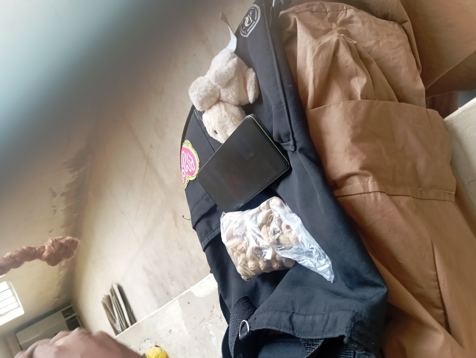 The image shows a close-up view of a black backpack with a brown jacket draped over it. Several items are placed on or near the backpack, including a smartphone, a small, light-colored plush toy, and a clear plastic bag filled with what appear to be nuts or seeds. The background is a wall with a rough texture, possibly concrete or plaster, with some discoloration and marks. There's a window with a light visible through it in the background, and a hint of some sort of shelving unit. The lighting suggests it could be daytime, but the overall scene is indoors and somewhat dimly lit. The angle of the shot is from above and to the side, looking down at the backpack and its contents. There is a partial view of a person's hand in the lower left corner of the image. Text is visible on a circular patch on the backpack, which appears to be a logo or emblem with some lettering, and also on a pink sticker with some illegible text. The image has a shallow depth of field, with the focus on the items on the backpack and the background being somewhat blurred.