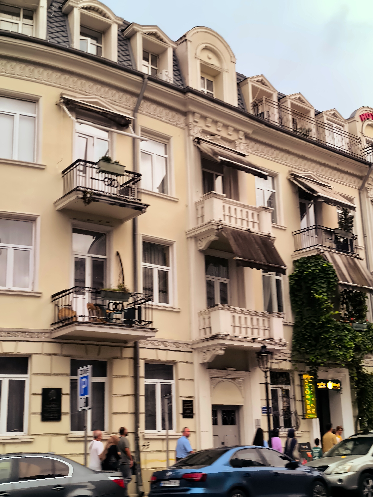 The image captures a street scene in Batumi, Georgia, featuring the facade of a cream-colored building with classical architectural elements, including decorative molding and arched dormer windows on the mansard roof. Several balconies with black railings adorn the building, some featuring planters with greenery. A prominent "HOTEL" sign in red neon letters is visible at the top right. The street level shows storefronts, one with a bright green neon sign. A parking sign indicating "ZONE P" is also present. Several people are walking on the sidewalk, and cars are parked and driving on the street. The overall impression is of an urban environment with a blend of historical architecture and modern city life.