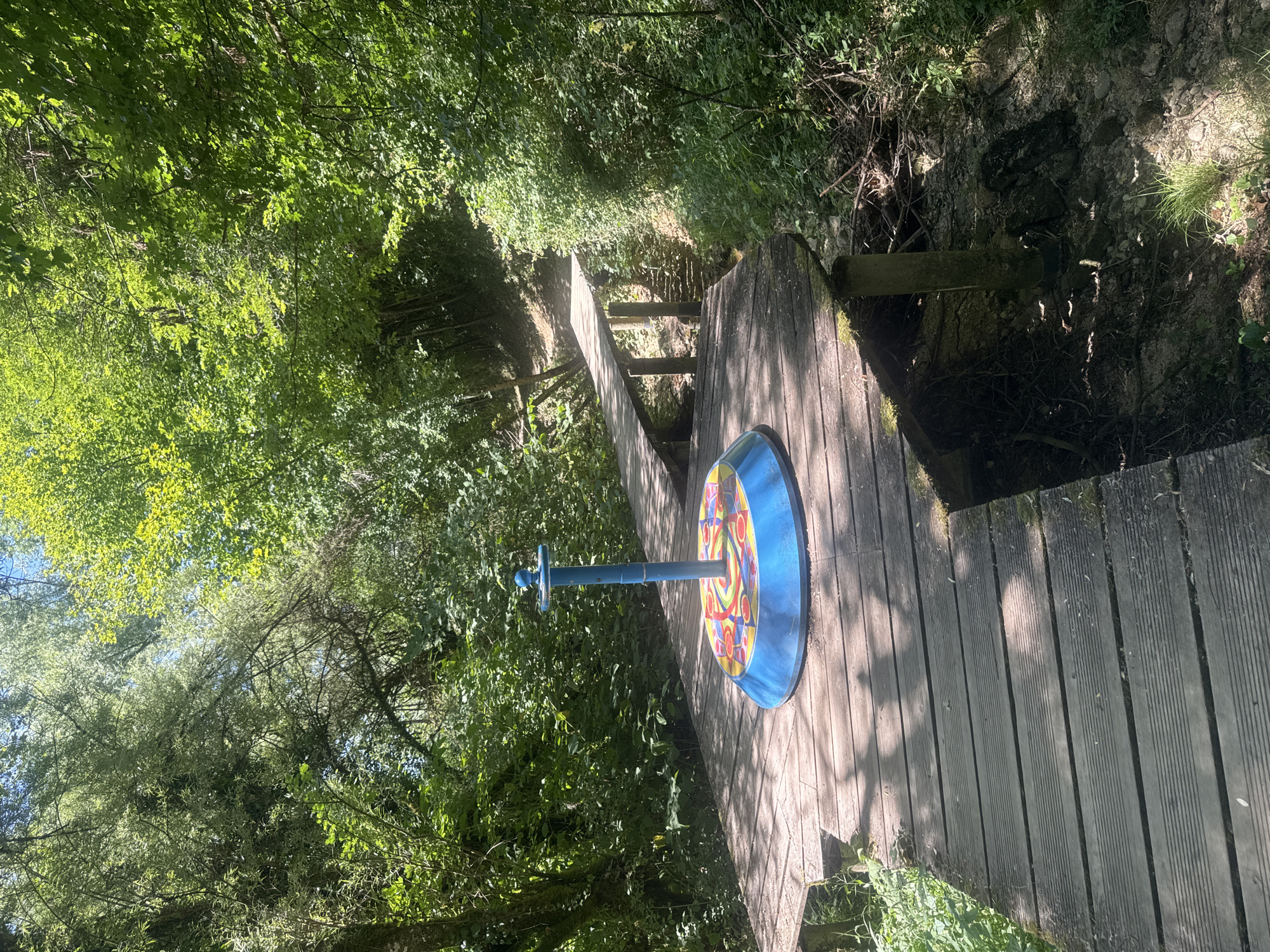 The image shows a colorful, circular merry-go-round situated on a wooden platform in a forest setting. The merry-go-round is blue with a brightly colored disc featuring patterns of red, yellow, and blue. A blue pole extends vertically from the center of the disc. The wooden platform is made of planks and appears to be part of a walkway or bridge in a wooded area. In the background, a second wooden bridge crosses over a small stream or ditch, leading uphill into denser foliage. The surrounding environment is lush and green, with sunlight filtering through the trees, casting dappled shadows on the platform. The location is identified as being in the city of Ravilloles, France.