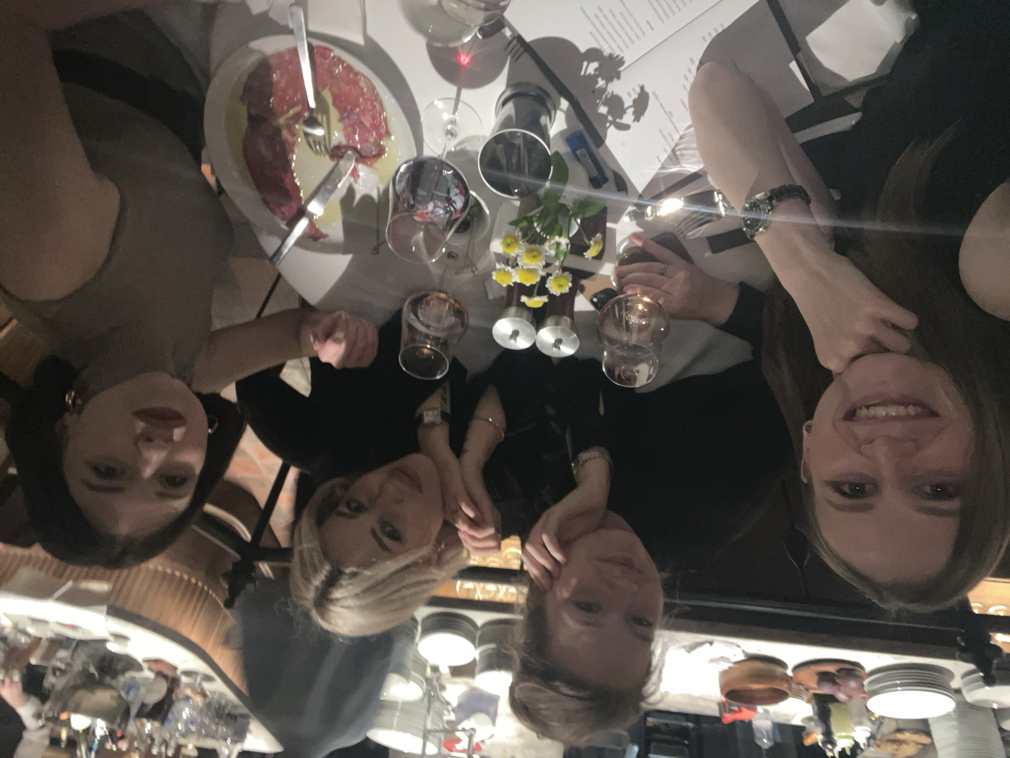Four women are gathered around a reflective dining table in a restaurant, possibly in Moscow, Russia, enjoying a meal or drinks during the evening. The image is taken from a top-down perspective, with the women's faces looking up towards the camera.
The subjects include four women. On the far left, a woman with dark hair and gold hoop earrings is visible. To her right, a blonde woman with a light-colored top is seen. Next to her, another woman with blonde or light brown hair and a dark top is present, with her hands clasped. On the far right, a woman with brown hair, smiling broadly, is visible. All women appear to be in good spirits, some smiling.
On the white table, several items are laid out. A plate of thinly sliced meat, possibly carpaccio, dressed with olive oil or sauce and garnished, is visible with a fork and knife. Several wine glasses, some appearing to contain red wine, are scattered across the table. A tall pepper mill stands near the center, alongside a small vase holding white and yellow daisy-like flowers. Small glass votive holders or decorative elements are also present. A menu with illegible text is open on the table, indicating a dining setting. The table's highly reflective surface mirrors parts of the women's faces, the restaurant's ceiling, and other ambient details, creating a shimmering effect.
The setting is an indoor restaurant with dim, warm lighting, suggesting a cozy evening ambiance. The activity is a social gathering, likely a dinner or drinks outing among friends. No specific weather conditions are discernible, as it is an indoor scene. The visible text on the menu is not legible.