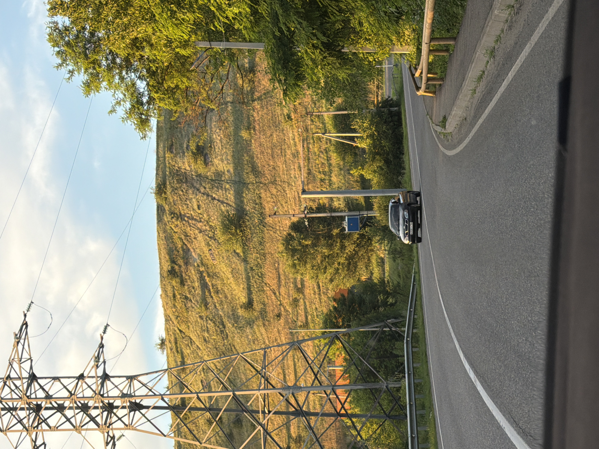 The image shows a rural road scene with a car driving on it. In the background, there is a large electrical tower, a grassy hill, and trees. The sky is blue with some clouds. A car is visible in the distance on the road. The road appears to be in a rural or semi-rural area. There are no people visible in the image. The image was taken from inside a vehicle, as suggested by the foreground which appears to be the dashboard. The scene is set during daylight hours, likely late afternoon or early evening given the golden hue of the sunlight on the hill. The environment is natural, with a focus on the landscape and infrastructure. The specific location is identified as Aleksandrovskoye, Russia.