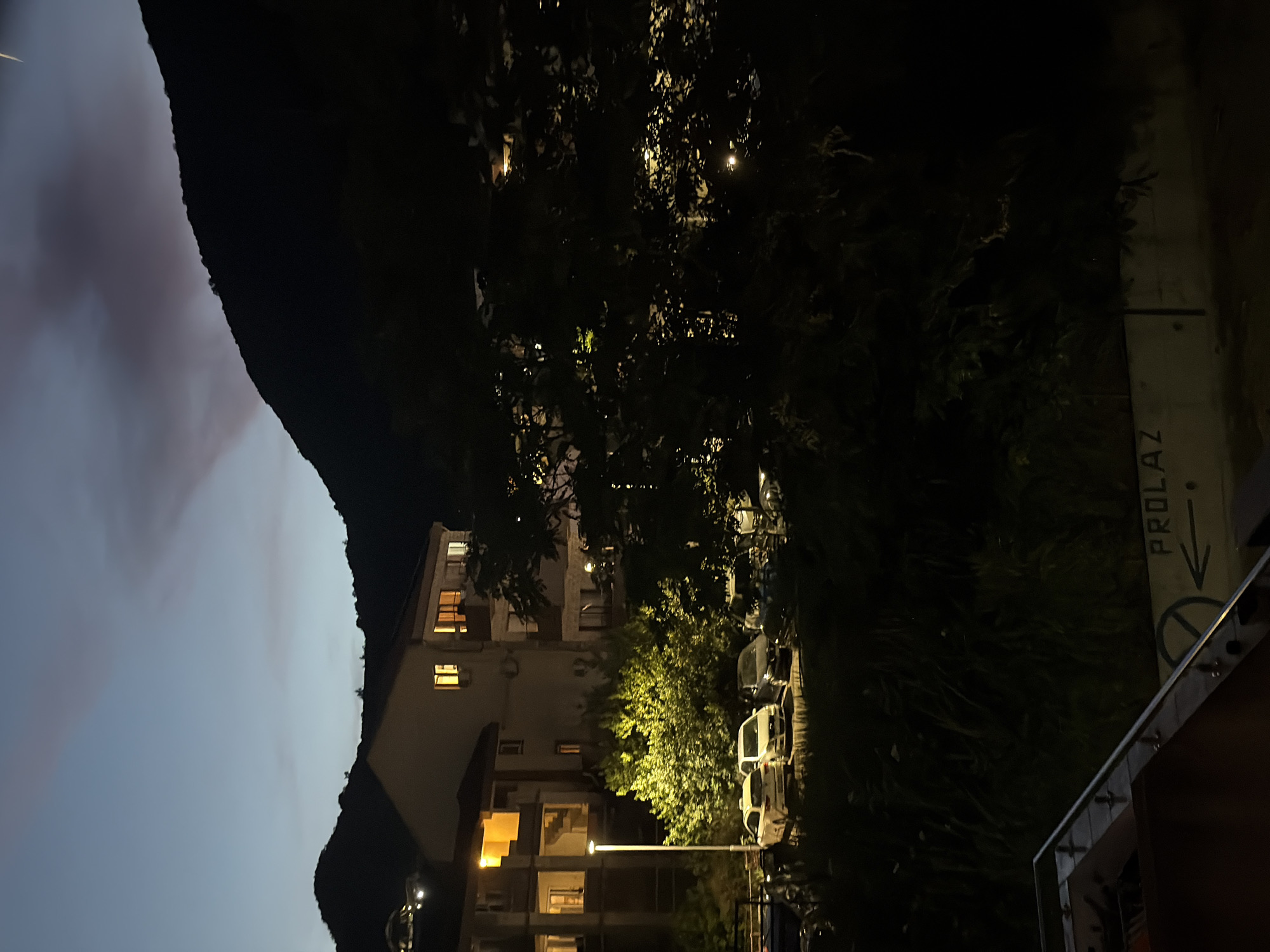 The image depicts a dusk scene in Budva, Montenegro, with a view of apartment buildings and a dark, tree-covered hillside. In the foreground, a metal railing of a balcony is visible. Below the balcony, a street or parking area is illuminated by a lamppost, with several cars parked. The apartment buildings have lit windows, suggesting inhabitants. Beyond the buildings and trees, the dark silhouette of a mountain rises against a twilight sky. The overall atmosphere is calm and nocturnal.