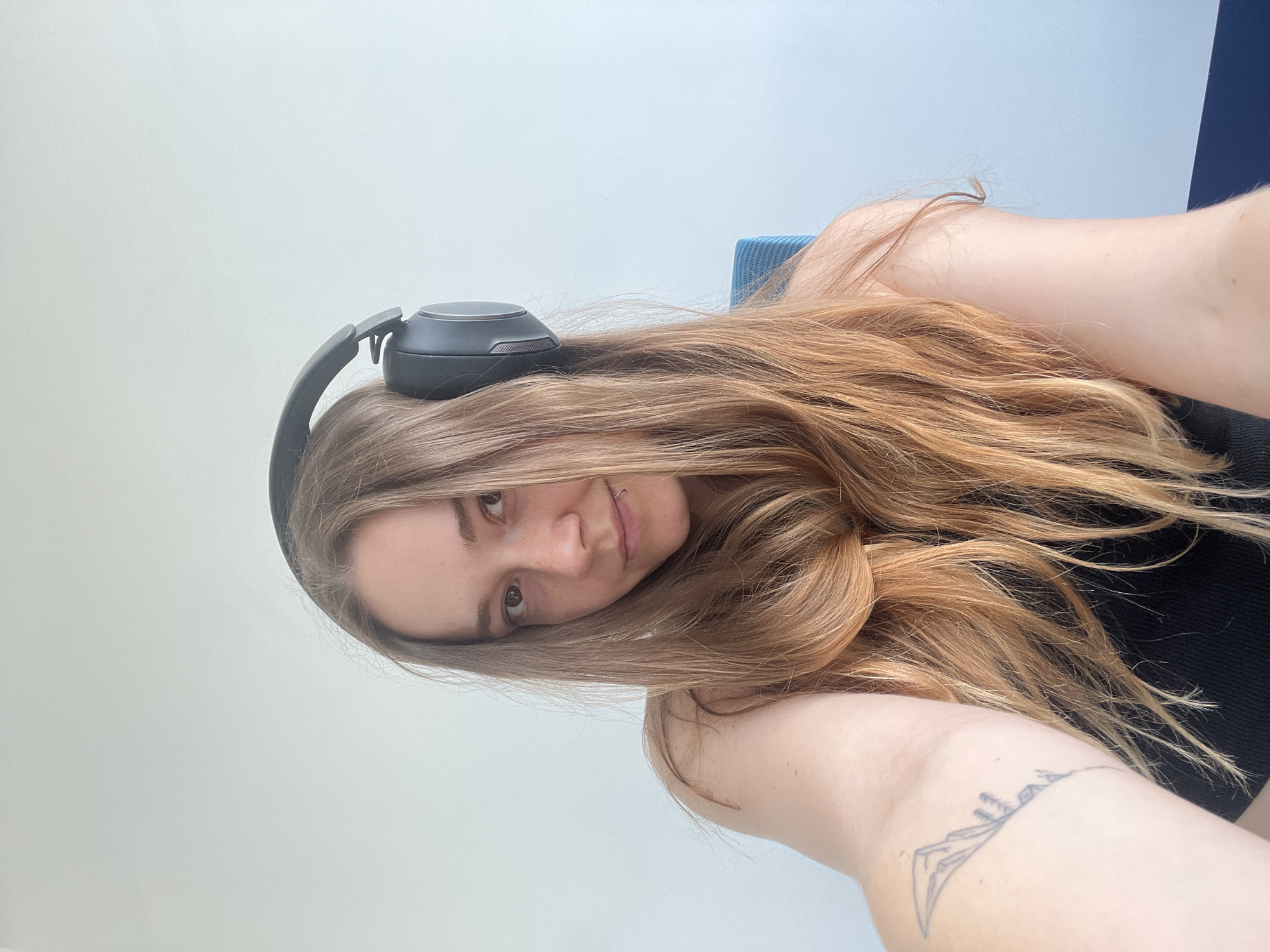 This media file is a selfie of a young woman with long, blonde hair, wearing black headphones. She is sitting in what appears to be an office or study, with a light-colored wall in the background and a hint of a blue chair. Her gaze is directed towards the camera, and she has a slight smile, conveying a relaxed and casual mood. A small, minimalist tattoo of a mountain range is visible on her upper arm. The lighting suggests it is daytime, and there are no strong indications of specific weather conditions or a distinct location within Plovdiv, Bulgaria, beyond a generic indoor setting. The overall impression is that of a personal moment captured during a period of work or leisure.