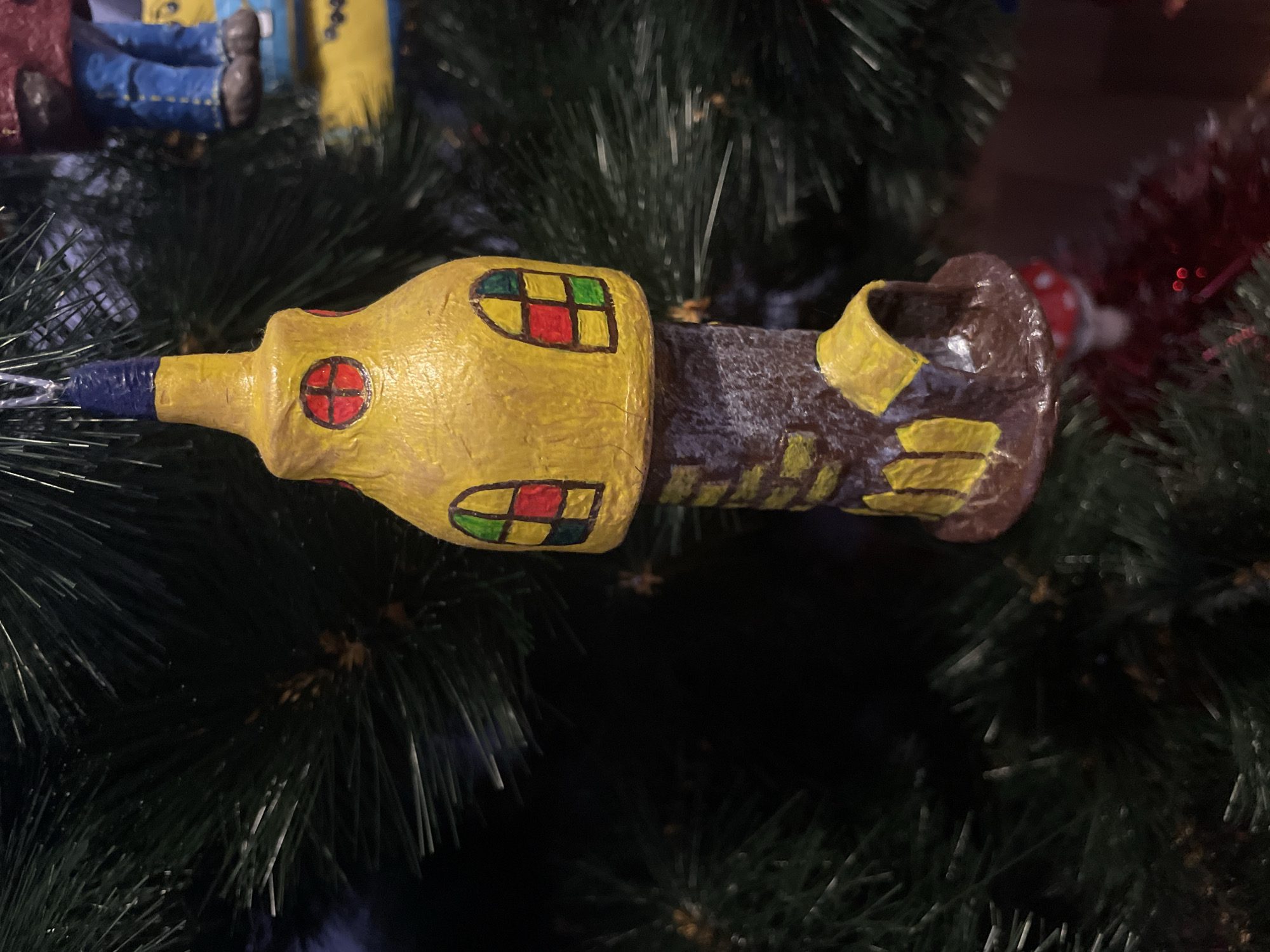 A handcrafted Christmas tree ornament, resembling a whimsical lighthouse or tower, hangs on a green artificial Christmas tree. The ornament is predominantly yellow and brown, with colorful window panes depicted in red, yellow, green, and blue. The top of the ornament is a pointed blue cap. The background is dark, with hints of other ornaments visible, including what appears to be blue jeans and brown shoes belonging to another ornament to the right. The scene is consistent with an indoor holiday decoration, likely in a residential setting in the city of Perm, Russia, given the context of the query. No people are visible in the frame.
