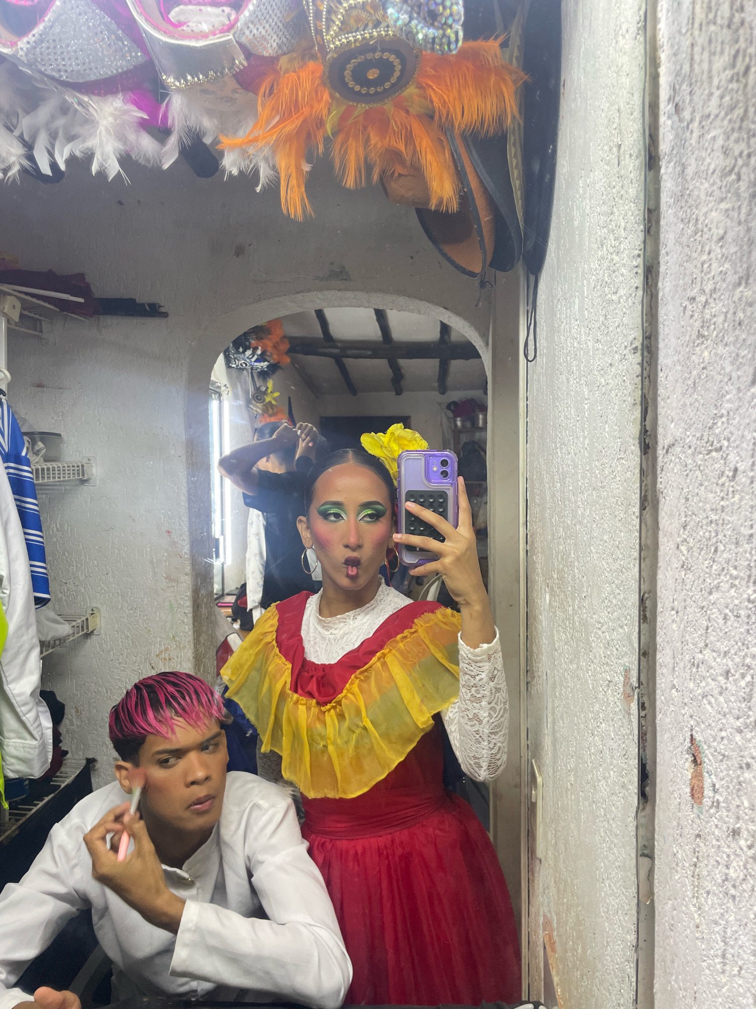 In a dimly lit dressing room, a young person with vibrant pink hair applies makeup to their face with a brush. They are wearing a white collared shirt. To their right, a young person in a colorful traditional dress, featuring a red skirt and a ruffled yellow and red bodice, poses for a selfie. Their face is adorned with dramatic green eye makeup and a yellow flower in their hair. In the background, another person's hands are visible, tending to hair. The room is cluttered with performance costumes and decorations hanging from the ceiling, including feathered headdresses and sequins. The walls appear to be made of a rough, textured material. The scene is captured through a mirror, reflecting the performers and their surroundings. The location is likely a backstage area associated with a cultural event or performance in Porlamar, Venezuela.
