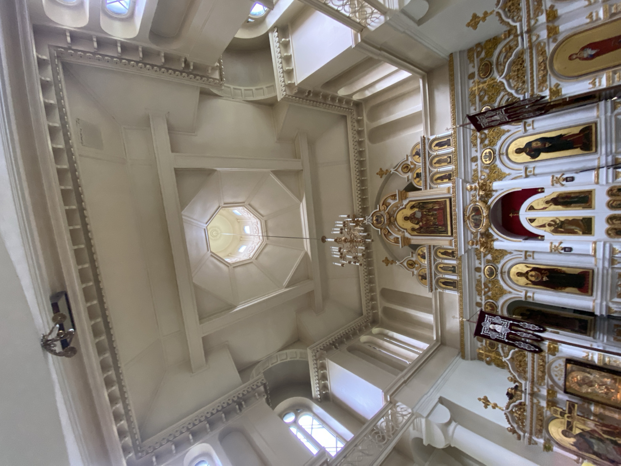The image captures the ornate interior of an Orthodox church, likely the Kazan Icon of the Mother of God Cathedral in Priozersk, Russia, based on its distinctive architectural features. The perspective is upward, emphasizing the grandeur of the ceiling and the iconostasis.
The ceiling is a complex architectural element, featuring a central octagonal dome with arched windows admitting natural light. Elaborate white molding and coffered details adorn the ceiling and walls, creating a sense of depth and grandeur. A large, multi-tiered chandelier with numerous candle-like lights hangs from the ceiling.
Dominating the lower half of the image is the iconostasis, a screen adorned with religious icons. The icons, depicting saints and biblical scenes, are framed by rich gold ornamentation and intricate carvings. Red banners hang from poles in front of the iconostasis, adding a splash of color to the scene. Arched doorways and niches are visible within the iconostasis, some of which contain smaller icons or are draped with red fabric.
The walls are painted a light color, likely white or cream, contrasting with the gold accents of the iconostasis. Tall, fluted columns support the upper architectural elements. Stained-glass windows, seen in the upper sections of the walls, contribute to the atmospheric lighting.
No people are visible in the frame. The scene is static, depicting the solemn beauty of the church interior. The overall impression is one of religious reverence and historical significance.