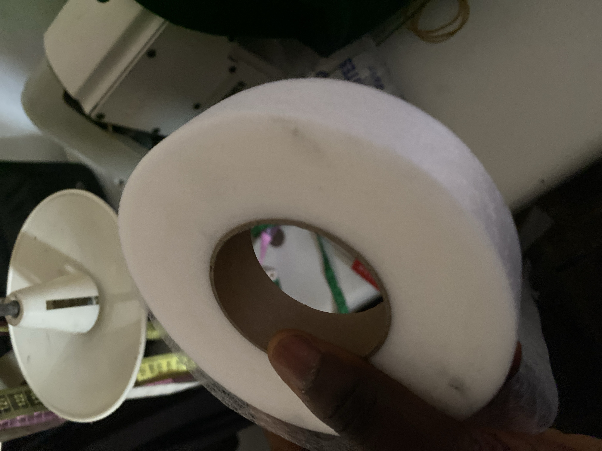 A person's hand with dark skin is holding a large roll of white fabric, possibly tulle or netting, with a cardboard tube in the center. The fabric is soft and appears to be a single, continuous piece wound around the tube. The background is blurry and appears to be an indoor setting, possibly a tailor's shop or craft room, with sewing machines and fabric scraps visible. There is a white, cone-shaped object with a metallic strip on its side in the upper left of the image, and a portion of a white sewing machine in the upper right. The lighting suggests an artificial light source. The scene is not identifiable as being in Jalingo, Nigeria.
