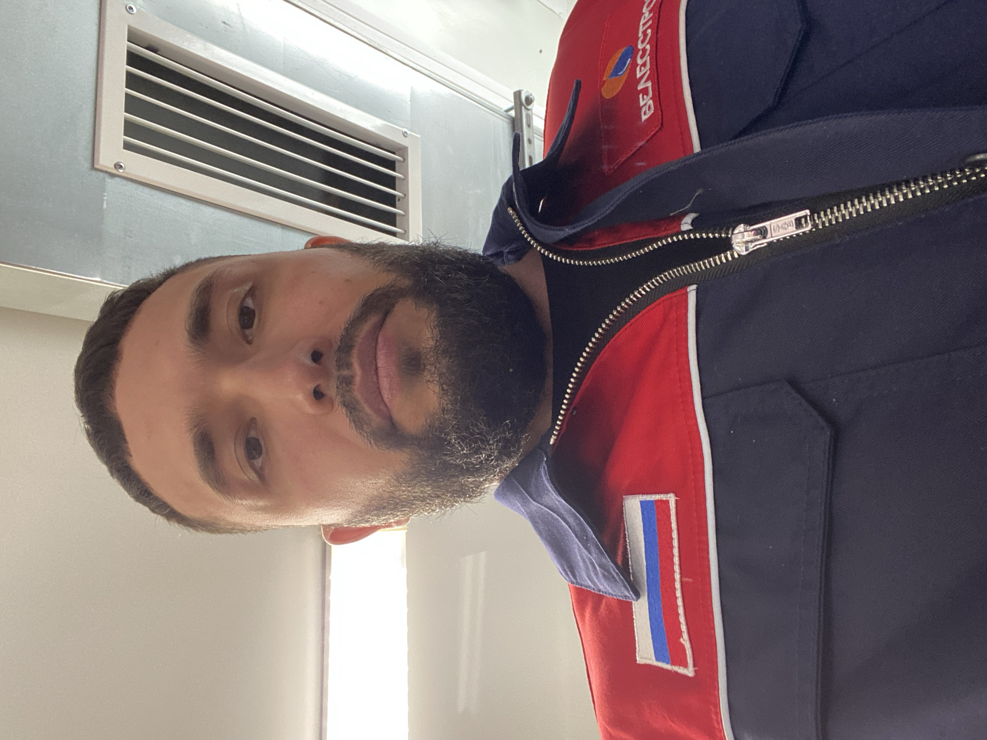 An indoor, portrait-style shot features a man with a dark beard and hair, looking directly at the camera. He is wearing a distinctive work jacket that is dark blue with a prominent red section on the chest and sleeves, separated by a thin white stripe. A silver zipper runs down the front, with the "YKK" brand name visible on the pull.
On the left side of his chest, there is a patch resembling the Russian flag with vertical white, blue, and red stripes. On the right chest area, a logo displays the Cyrillic text "БЕЛЭНЕРГОСТРОЙ" (BELENERGOSTROY) alongside a stylized design incorporating red, orange, and blue elements.
The background consists of a light-colored wall or ceiling, with a rectangular metal ventilation grate mounted high on the surface. The lighting appears artificial, suggesting an indoor setting within an industrial or utilitarian facility in Luzhitsy, Russia. The man's expression is neutral, and his head is slightly tilted as he poses for the image.