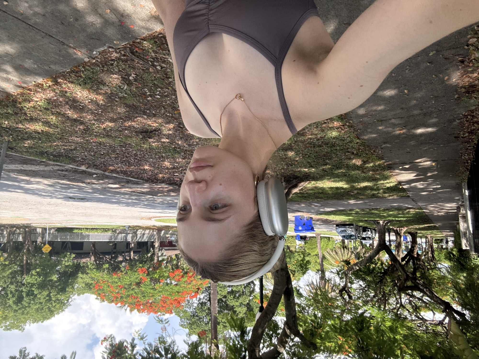 The image depicts a young Caucasian woman with blonde hair, wearing large silver headphones, a grey tank top, and a gold necklace. She is standing on a sidewalk in what appears to be a suburban residential area. The background shows a street with parked cars, trees with lush green foliage and some with bright orange flowers, houses with white fences, and a blue recycling bin. The presence of palm trees and the style of houses suggests a location in Florida. The lighting indicates it is daytime, with shadows cast by the trees. The overall scene is one of a casual outdoor setting, likely in a park or along a residential street. Based on the vegetation and architecture, this scene is consistent with a location in Miami, Florida, United States of America.