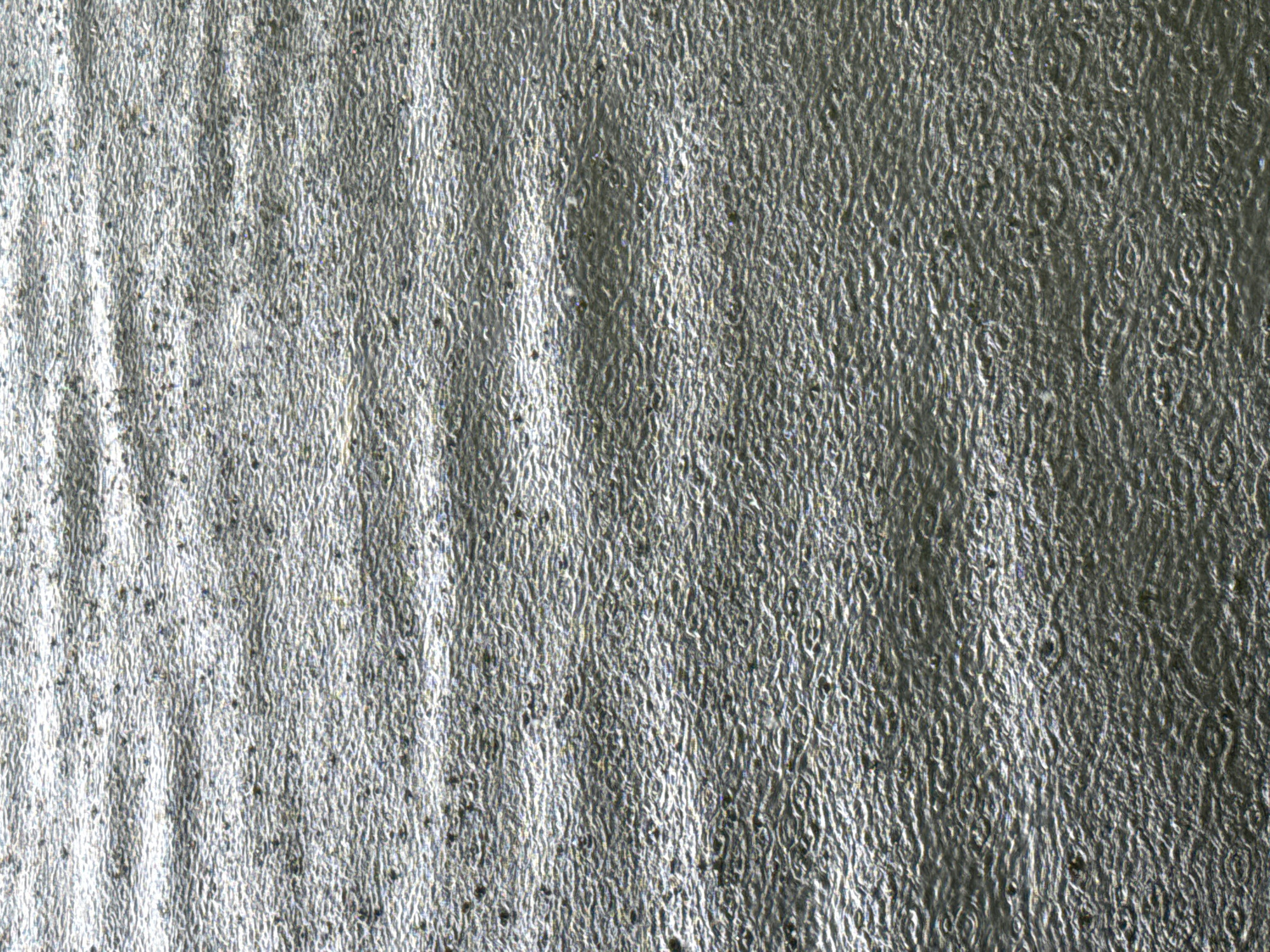 The image shows a close-up, abstract view of a textured, silvery surface that appears to be rippled or wave-like. The texture is consistent across the entire frame, with horizontal bands of lighter and darker shades suggesting a repeating pattern or the play of light on the surface. Small, dark specks are scattered throughout the texture, adding to its granular appearance. The lighting creates highlights and shadows that emphasize the undulations of the surface. This image does not contain any discernible elements of a city, people, or specific actions, and therefore cannot be localized to Huizhou, China, or any other specific geographic location.