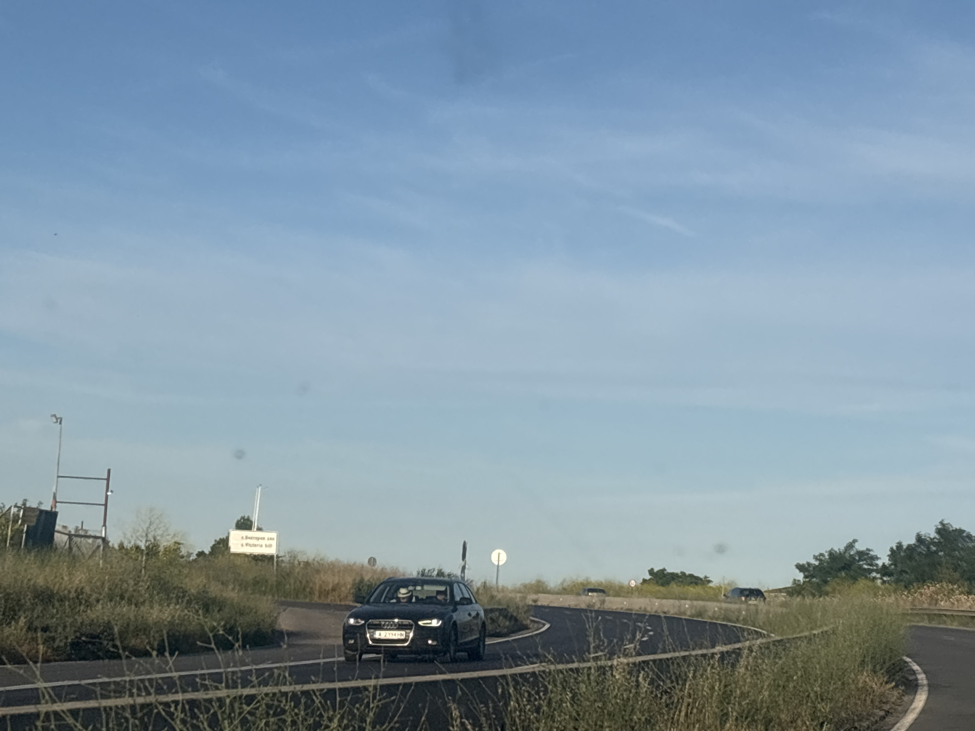 The image shows a road in Pomorie, Bulgaria. A black Audi A4 is driving on the road, with two people inside. The car is facing the camera. There is a signpost in the background, and some trees and grass on the side of the road. The sky is blue with some clouds. The scene appears to be a sunny day.