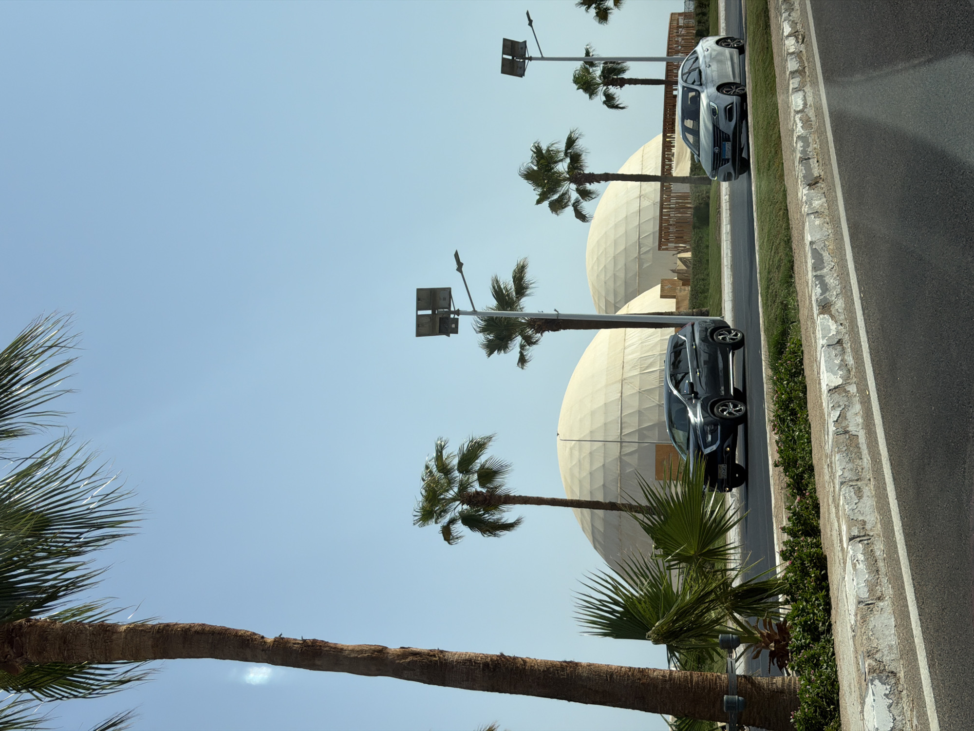 The image shows a street scene in Sharm El Sheikh, Egypt, with a clear blue sky and several palm trees framing the view. In the background, two large, white, dome-shaped structures are visible. These are likely part of a venue or exhibition center. A dark-colored SUV is driving in the left lane, and a silver sedan is in the right lane. The road has white markings and a landscaped median with grass and some flowering plants. The foreground shows a portion of the asphalt road and a concrete curb. There are streetlights visible on poles along the road. The overall impression is a bright, sunny day in a coastal city.