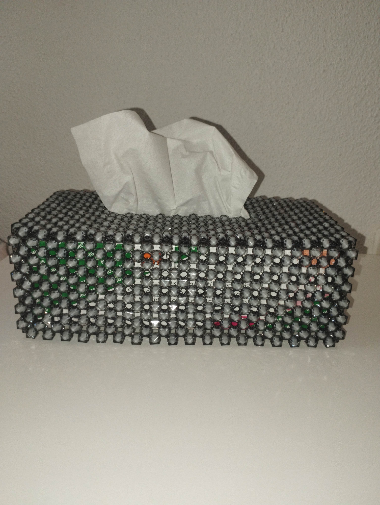 The image displays a rectangular tissue box, covered with an ornate, handcrafted beaded casing, resting on a flat, light-colored surface. A few crumpled white tissues are visibly protruding from the opening at the top of the box.
The decorative cover of the tissue box is made from numerous translucent, faceted beads, appearing dark gray or black in color. These beads are arranged in a hexagonal, honeycomb-like pattern, creating a mesh through which the original packaging of the tissue box is partly visible. The underlying tissue box packaging features various colors, including prominent green sections, as well as hints of orange, red, and white.
Through the gaps in the beaded cover, fragmented and partially obscured text is discernible on the tissue box packaging. On the left side, letters like "RO", "IG", and "ri" can be seen. On the front surface, isolated letters and partial words such as "tic", "en", "his", "K", "I", "d", and "c" are faintly visible, making it difficult to decipher complete words or phrases.
The object is placed against a light, off-white or beige wall with a subtly textured finish. The lighting suggests an indoor setting, likely under artificial illumination, as there are no strong shadows indicative of natural daylight. There are no people present, and no specific activity is taking place; the image is a static shot of the item. The overall impression is one of a functional household item given a decorative, personalized touch.