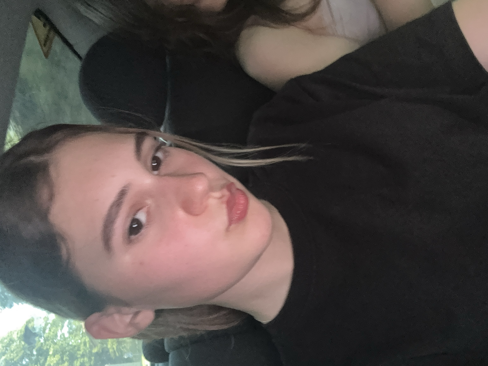 The media file shows two people in a car, with a focus on the face of a young woman in the foreground. She has light brown hair with blonde streaks, which are falling across her face. Her eyes are dark brown and she is looking directly at the camera. Her lips are slightly parted and appear to have a pink or red tint. She is wearing a dark t-shirt. Behind her, another person is partially visible, also wearing dark clothing. The interior of the car is dark, with a hint of greenery and trees visible through the window, suggesting an outdoor setting during the daytime, possibly a road trip. The lighting suggests it might be late afternoon or early evening. The overall mood appears casual and intimate, possibly a selfie taken by the young woman. There is no visible text in the image.