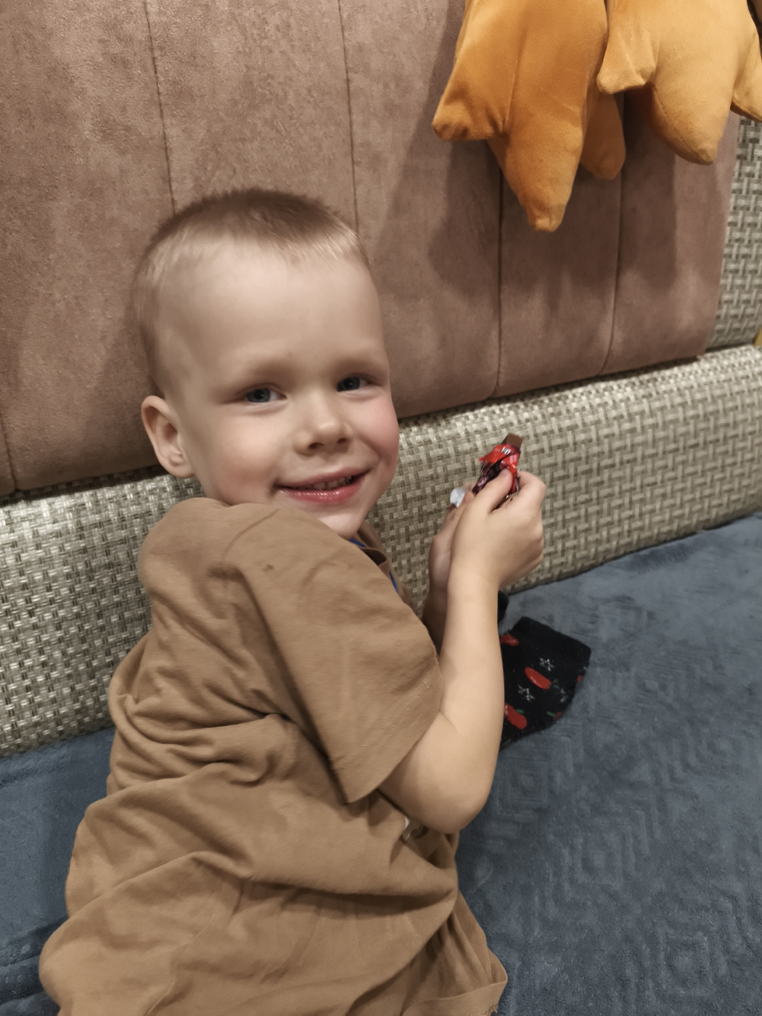 A young boy is lying on a gray surface, likely a bed or sofa, with his head near a beige, textured upholstered backrest. A tan colored, short-sleeved shirt covers his upper body. He is holding a small, wrapped candy bar in his hands, smiling broadly, displaying his teeth. A single black sock with a pattern of red shapes is visible near his hand. An orange plush object is also visible, partially in frame, at the upper right of the image. The location is Русский Вожой, Russia.