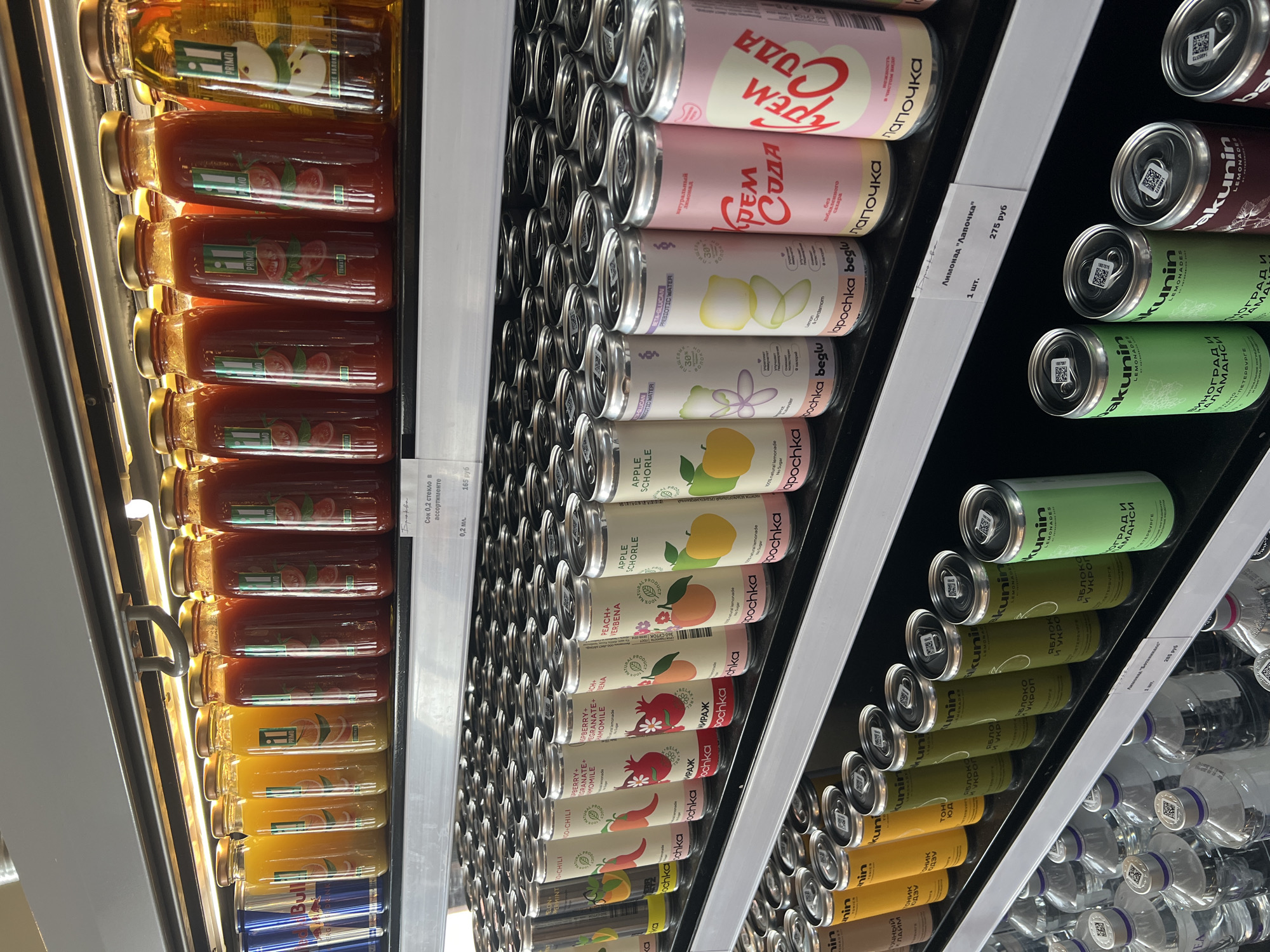 The image shows shelves in a refrigerated display case filled with various beverages. On the left are glass bottles of juice, including apple, tomato, and orange flavors.
The central section showcases cans of "Lapochka" (Лапочка) soda in different flavors, such as cream soda and apple schorle.
On the right are cans of "Bakunin" (Bakunin) lemonade, including flavors like apple and dill, and grape and basil.
Price tags are visible for both the "Lapochka" and "Bakunin" lemonades, indicating a price of 275 rubles and 285 rubles, respectively. The shelves also hold bottles of other drinks, including Red Bull and sparkling water. The "Bakunin" cans state that they are produced in Saint Petersburg (Санкт-Петербург).