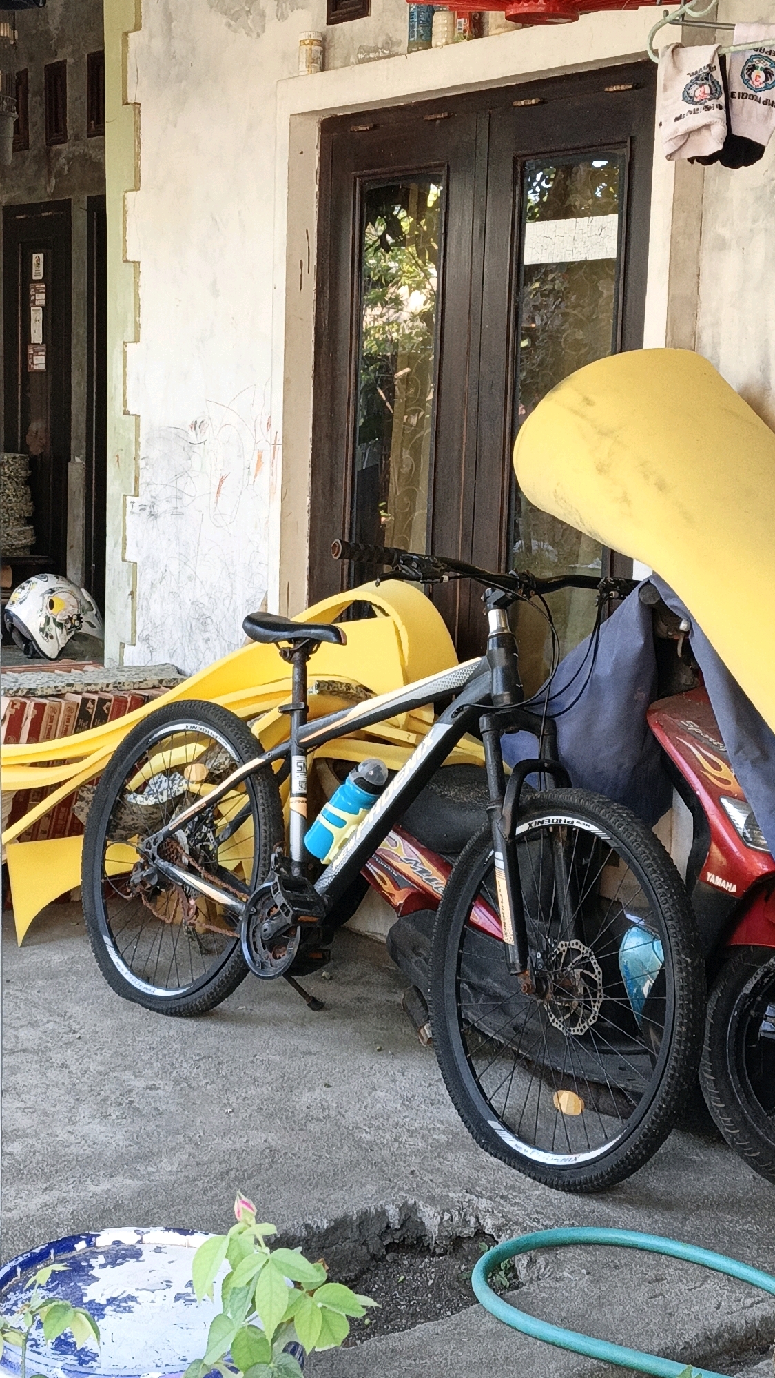 A black mountain bike is parked on a concrete surface in front of a building with brown double doors and a light-colored textured wall. The bike has a blue water bottle attached to its frame and a yellow object leaning against it. To the right of the bike, a red motorcycle is visible. In the background, there are some items hanging on a clothesline. The scene appears to be outdoors, possibly in a residential or commercial area. The image is taken from a slightly low angle, looking up towards the bike and the building.