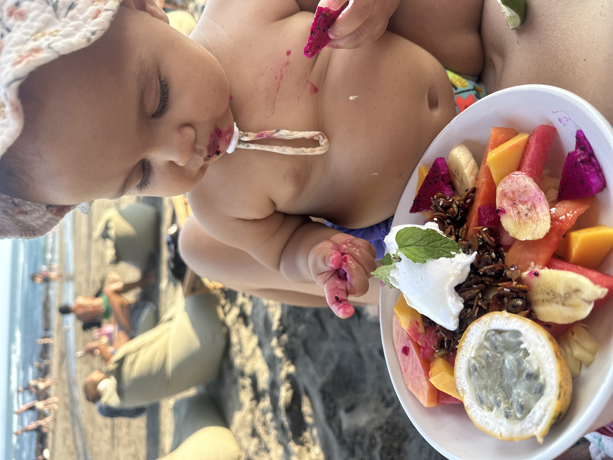An overhead shot shows a toddler sitting on the sand on a beach in North Kuta, Indonesia, eating a bowl of fruit. The toddler wears a floral bucket hat and a pink stain surrounds their mouth and chin, indicating they have been eating berries. Their right hand, stained pink, holds a piece of dragon fruit. A white bowl of fruit is in front of them, containing granola, yogurt, passion fruit, papaya, watermelon, banana, and dragon fruit. In the background, out of focus, are other beachgoers sitting on beanbag chairs and wading in the ocean. The sun casts a warm glow over the scene.