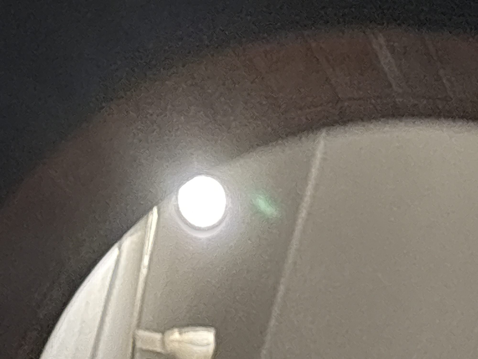 The image shows a close-up, slightly out-of-focus view of a light source, likely a ceiling fixture, emitting a bright white light. The light is circular and positioned towards the center of the frame. Below the light, there is a faint green lens flare. The surrounding environment appears to be an indoor space with neutral-colored walls or ceilings, possibly textured. A dark, curved element, potentially a structural beam or architectural feature, frames the upper right portion of the image. The overall impression is one of an internal, possibly residential or commercial, setting with a prominent artificial light source. This image is not specific to Ćwiklice, Poland, and does not contain any identifiable landmarks or details to suggest that specific location.