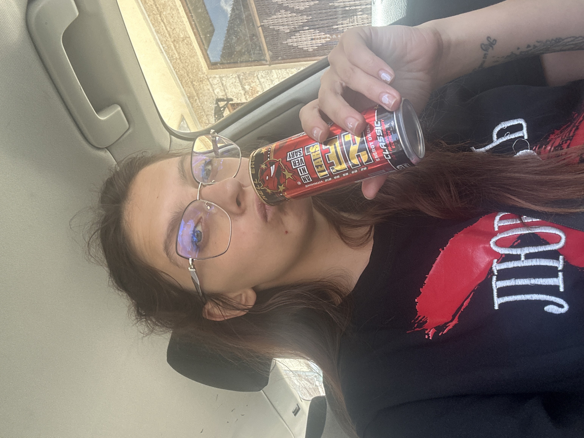 A young woman with long brown hair and glasses is in the passenger seat of a vehicle. She is holding a can of Hell Energy drink to her mouth and appears to be drinking it. She is wearing a black t-shirt with a red design and white lettering. The background shows the interior of a car and a brick building outside the window. The scene is likely in the city of Tryavna, Bulgaria, given the context provided.