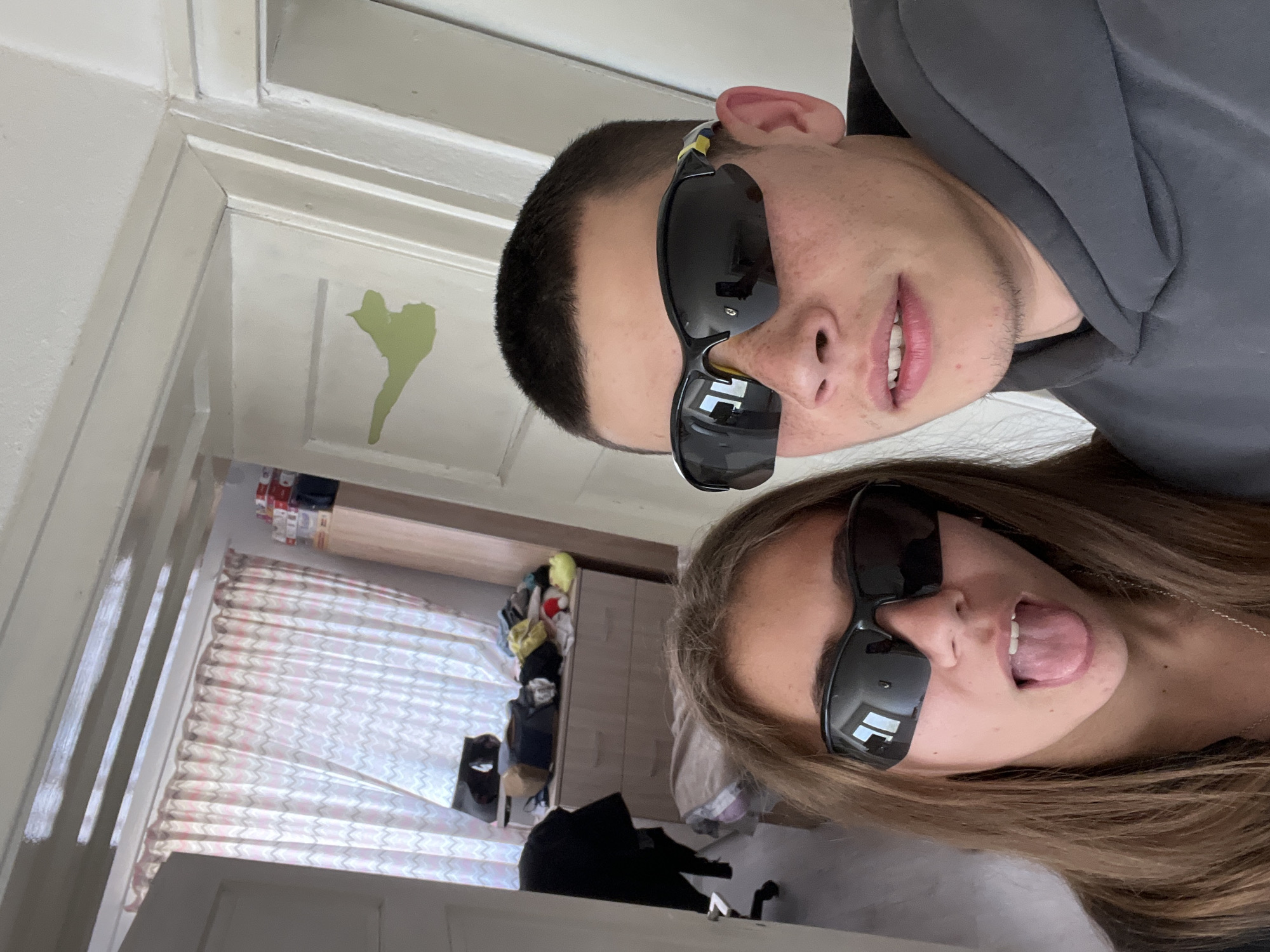 Two people are posing for a selfie indoors. The person at the top of the image is wearing sunglasses and a gray hoodie. The person at the bottom of the image is also wearing sunglasses and has their tongue sticking out. The background appears to be a bedroom with curtains and a closet. There is a green shape on a wall near a door frame. The location is unknown and there is no indication of the image being in Karavelovo, Bulgaria.