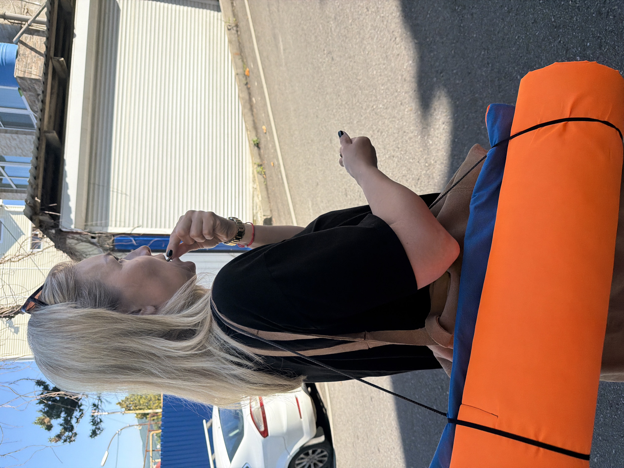 A middle-aged Caucasian woman with blonde hair tied back and sunglasses perched on her head stands outdoors, facing right in profile. She is wearing a black t-shirt and a brown skirt. The woman is bringing a small object to her mouth with her right hand and holding a rolled-up orange and blue mat with straps over her left shoulder. A white car is parked behind her on the left. The background consists of a building with a white metal corrugated door and some scaffolding. The scene is set on a paved street under a clear blue sky. This image was taken in the city of Sochi, Russia.