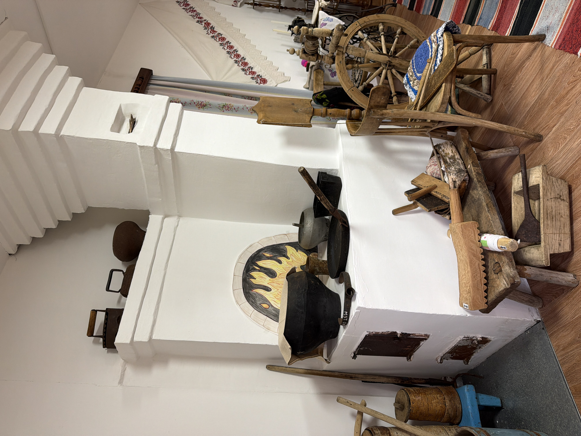 The image displays a rustic, indoor scene, likely a museum exhibit or a historical reenactment space, depicting traditional Russian village life. A large, white plaster stove dominates the center of the frame. On top of the stove, various antique household items are arranged, including two cast-iron clothes irons, a dark-colored pot, and a smaller metal pot.
In front of the stove, on a raised platform, sits a large, black cast-iron cauldron covered with a piece of fabric. Next to it are smaller metal pots and a frying pan. The front of the stove has two dark metal doors, likely for the oven and ashpit, with one visible handle. Above these doors, an arched opening in the stove is decorated with a painted depiction of flames.
To the left of the stove, several wooden objects are leaning against the wall, including long poles and a cylindrical wooden container with a lid, resting on a small blue stool.
To the right of the stove, a rustic wooden spinning wheel with a large wheel is prominently featured. It is placed behind a wooden chair with a woven mat on its seat. A stack of wooden tools and materials, including a flax hackle and a basket containing yarn, rests on a low wooden bench in front of the chair. Below the bench, a wooden trough holds more tools, including a long-handled shovel.
The floor is covered with polished wood, and a colorful striped rug runs along the right side of the frame. A white embroidered cloth with red floral patterns hangs on the wall in the background. The overall ambiance is one of historical preservation and cultural display.
While the image is identified as being located in the city of Ust'-Pyzas, Russia, there are no people visible in the scene. The objects are static and arranged for display. The environment appears to be indoors, with artificial lighting.